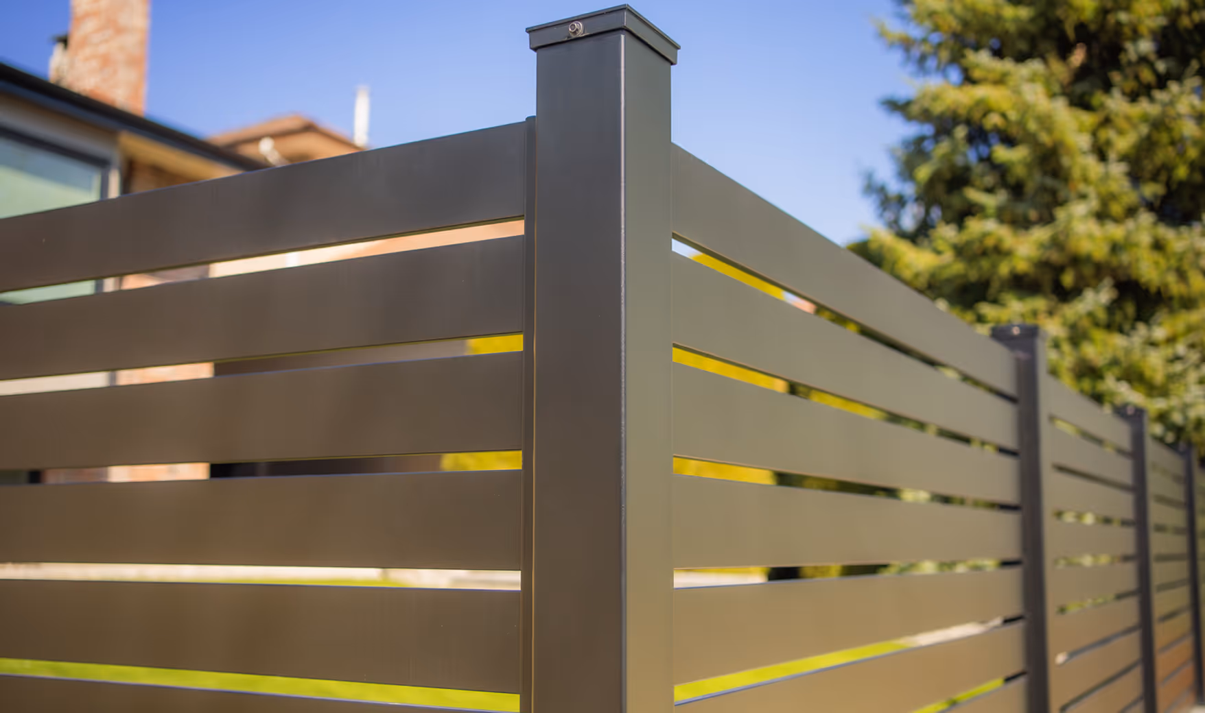 Modern horizontal slat fence made of dark metal panels in a residential area with trees and houses in the background.