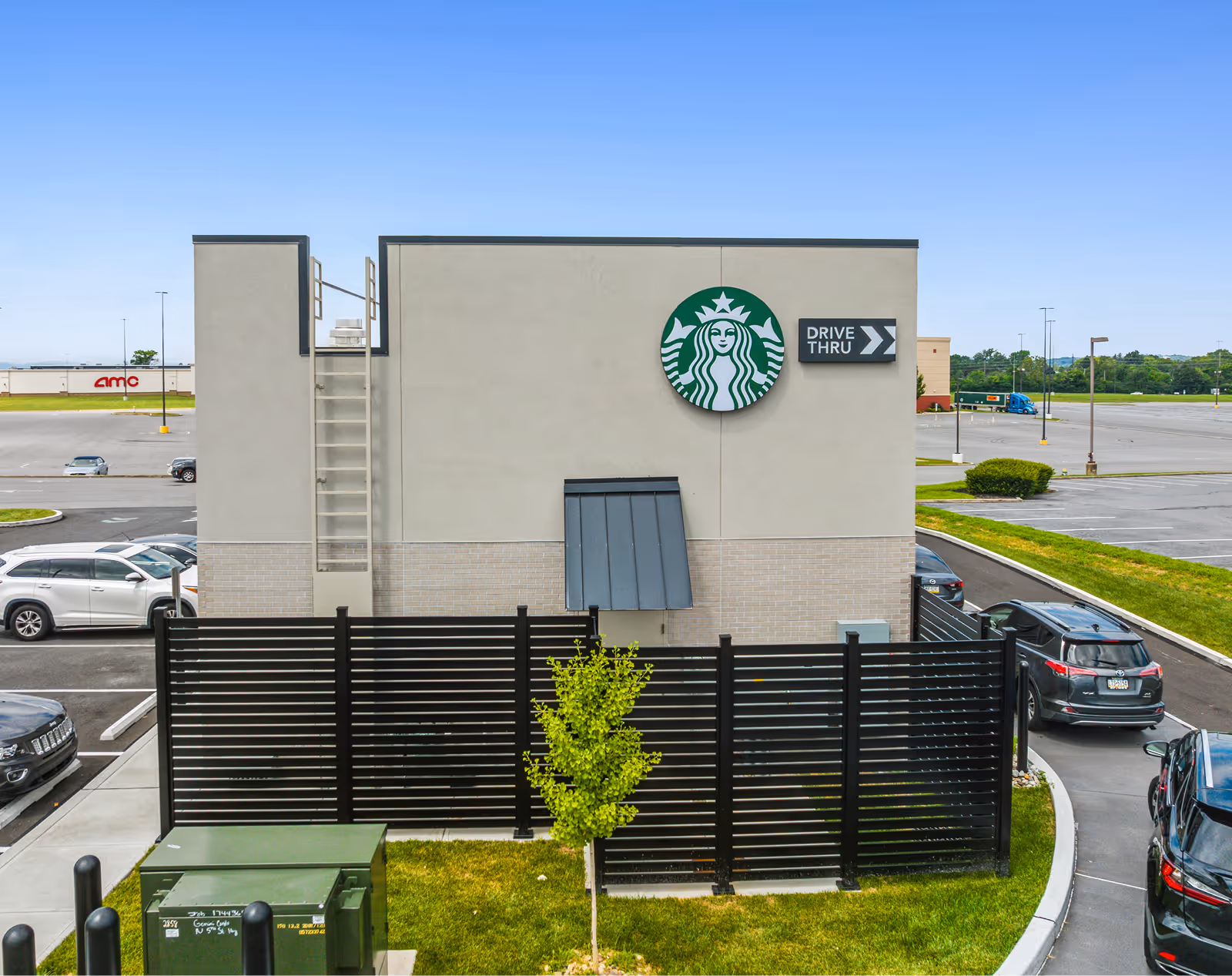 Exterior rear view of a Starbucks building with drive-thru signage and black horizontal slat fencing enclosing a small tree.
