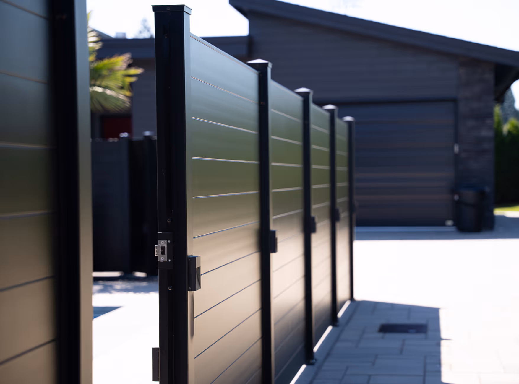 Modern black metal vehicle gate with horizontal panels partially open on a paved driveway in front of a garage.