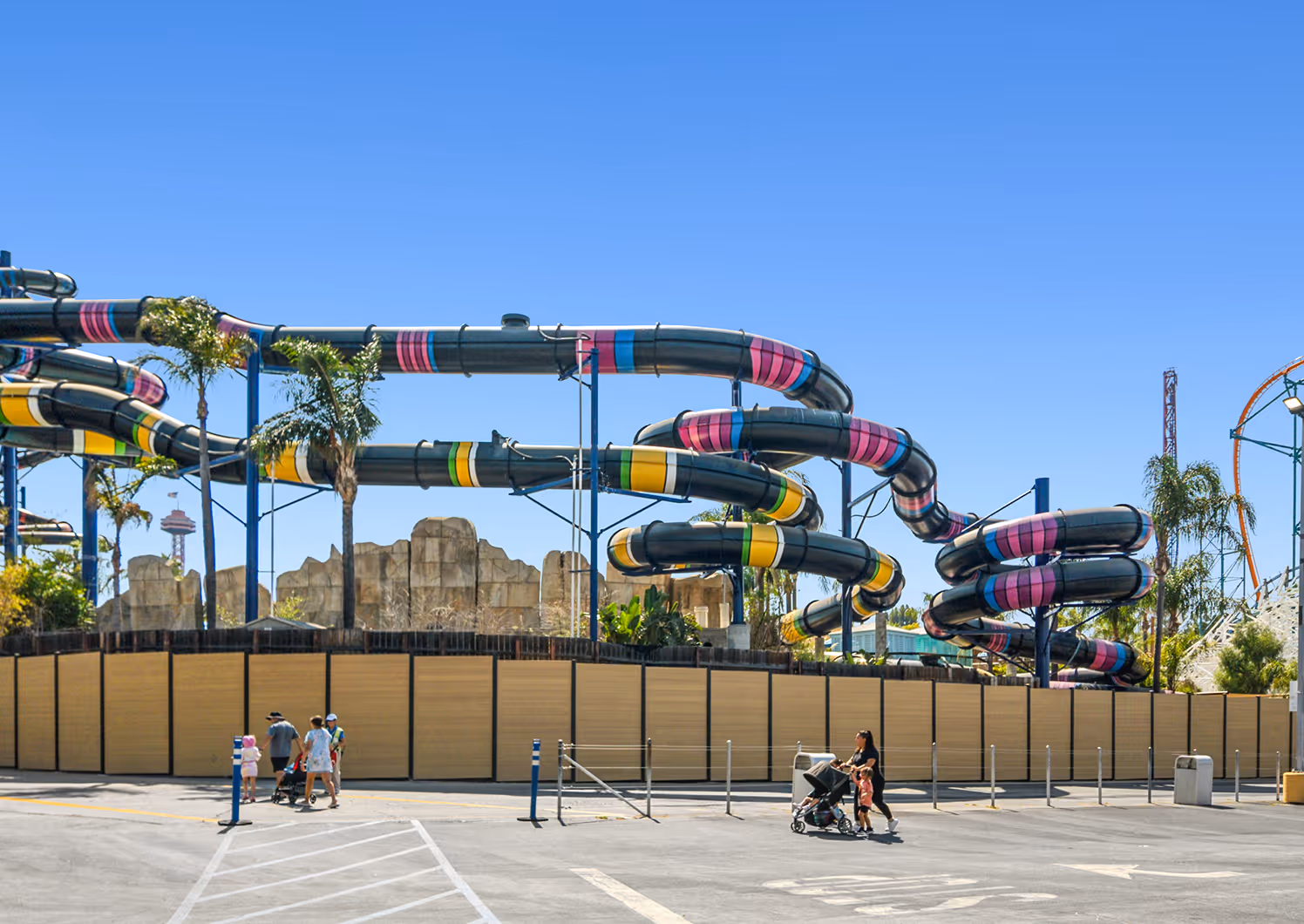 Curved colorful water slides at an amusement park with people walking and pushing strollers in the foreground on a sunny day.