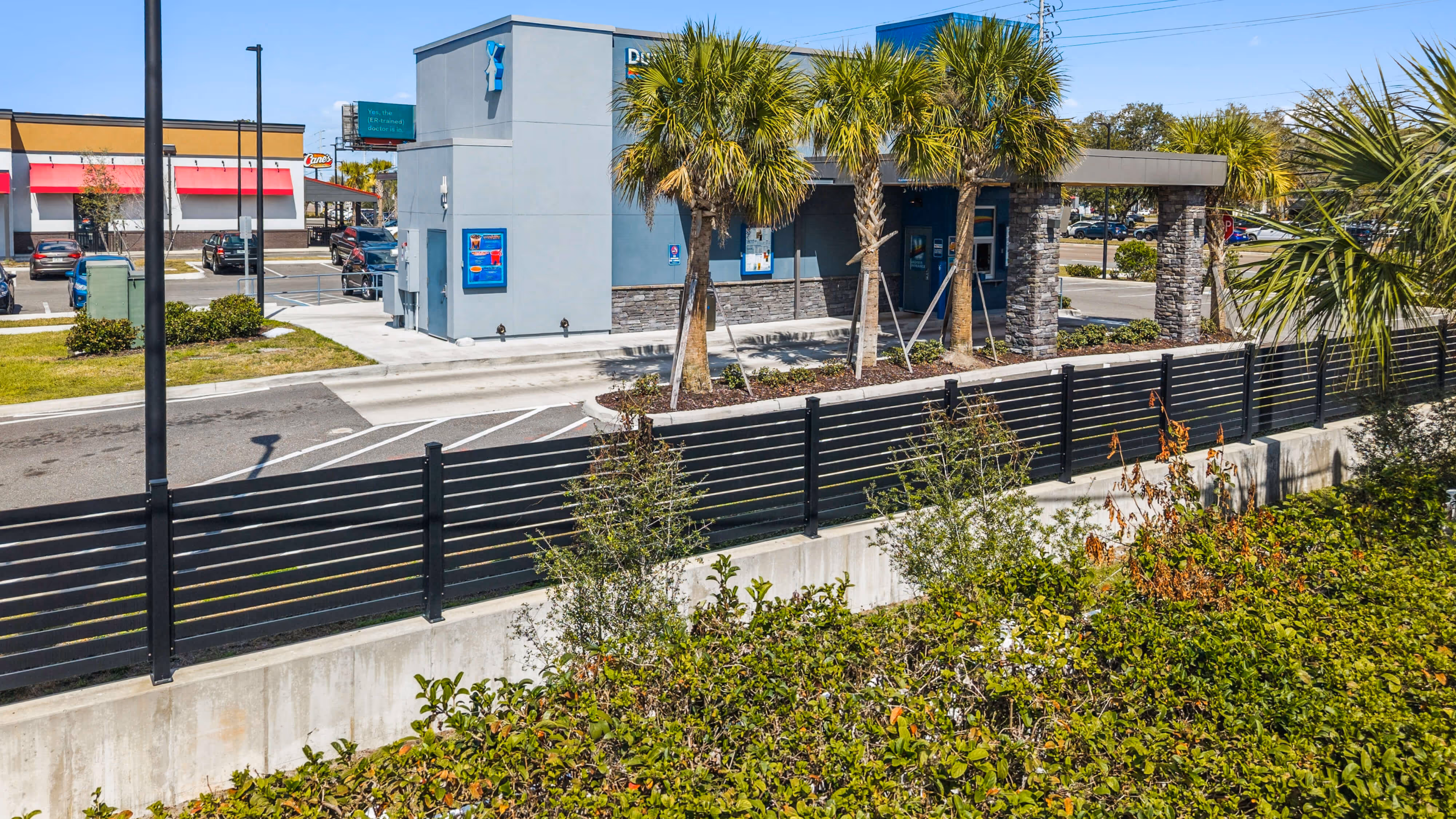 Black horizontal slat aluminum fence bordering a commercial drive-thru area with palm trees and shrubs.