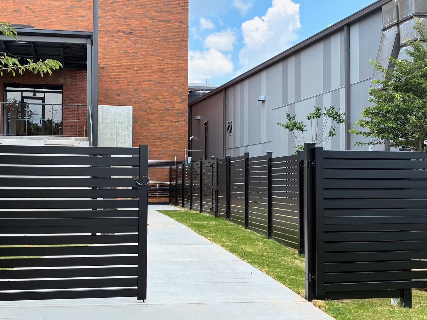 Black horizontal slat aluminum fence along a concrete pathway between brick and metal buildings with some greenery.