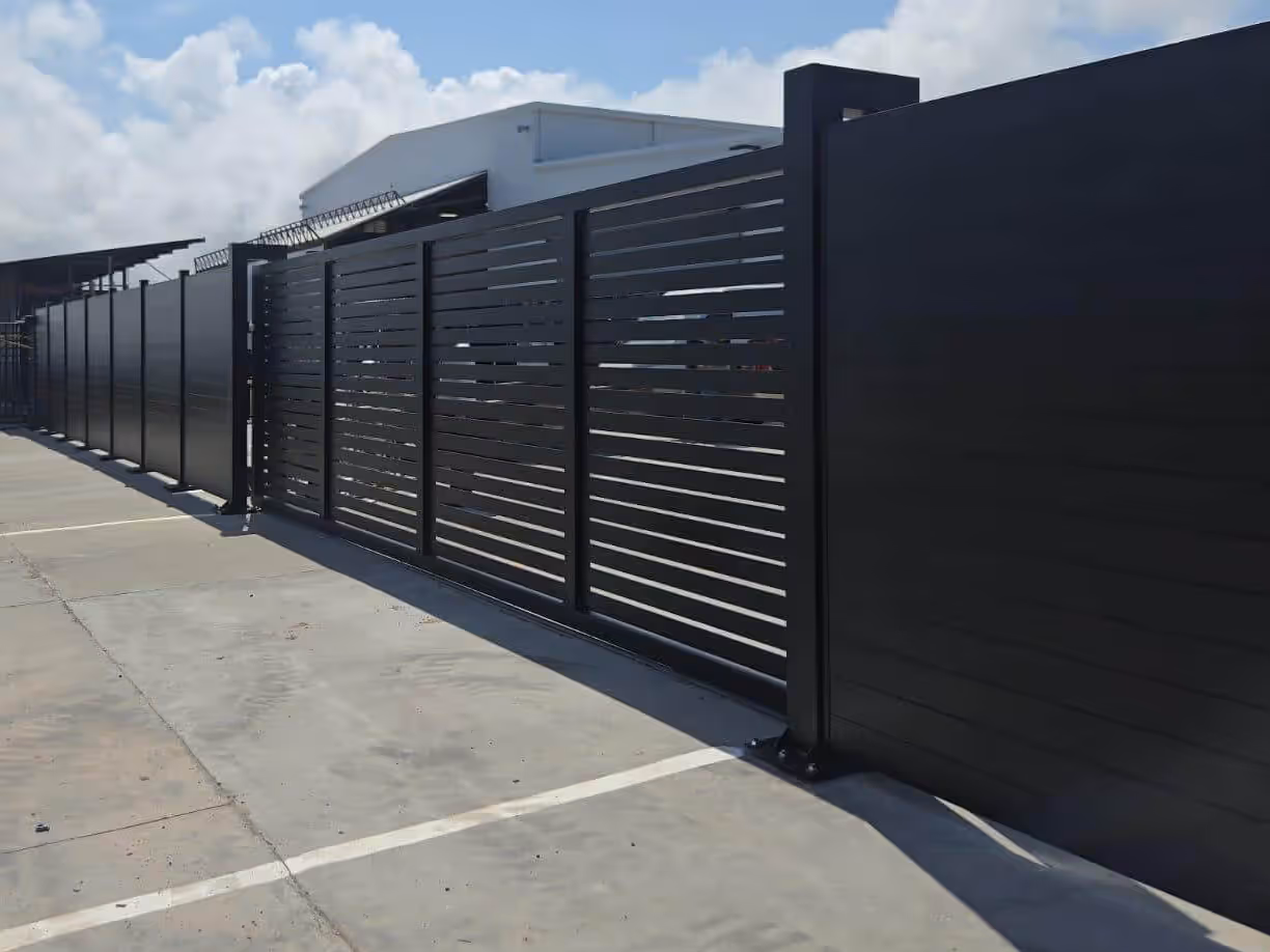 Long black horizontal slat metal driveway gate and fence on a concrete driveway with a building in the background.