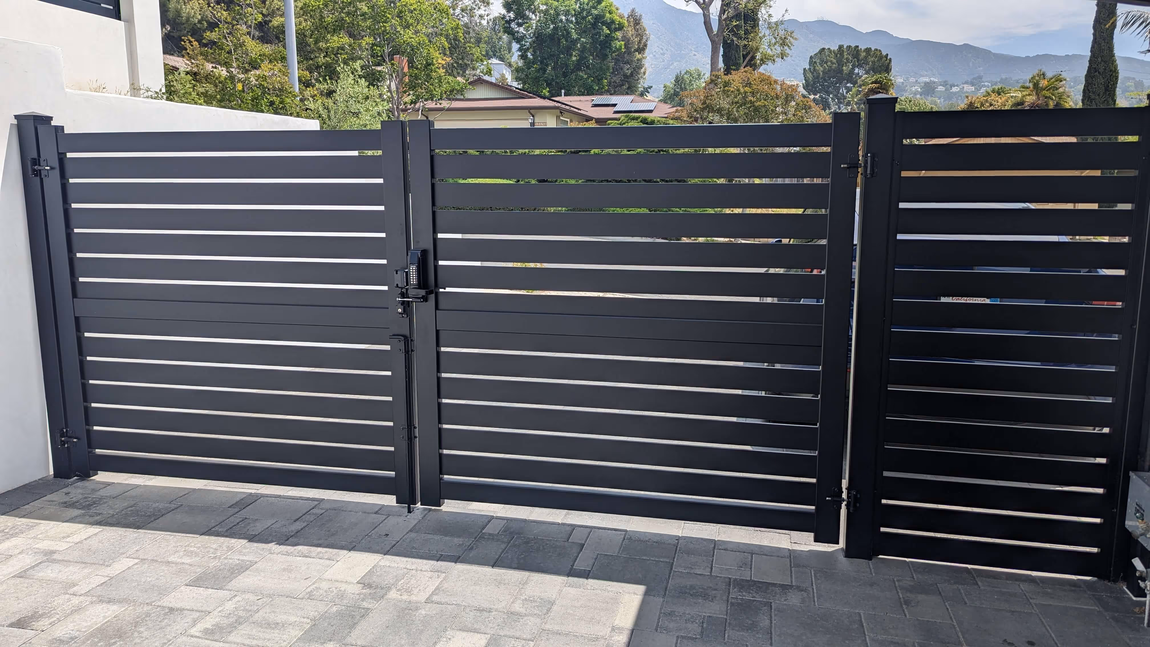 Modern black aluminum double swing gate with horizontal slats and keypad lock in a residential driveway.