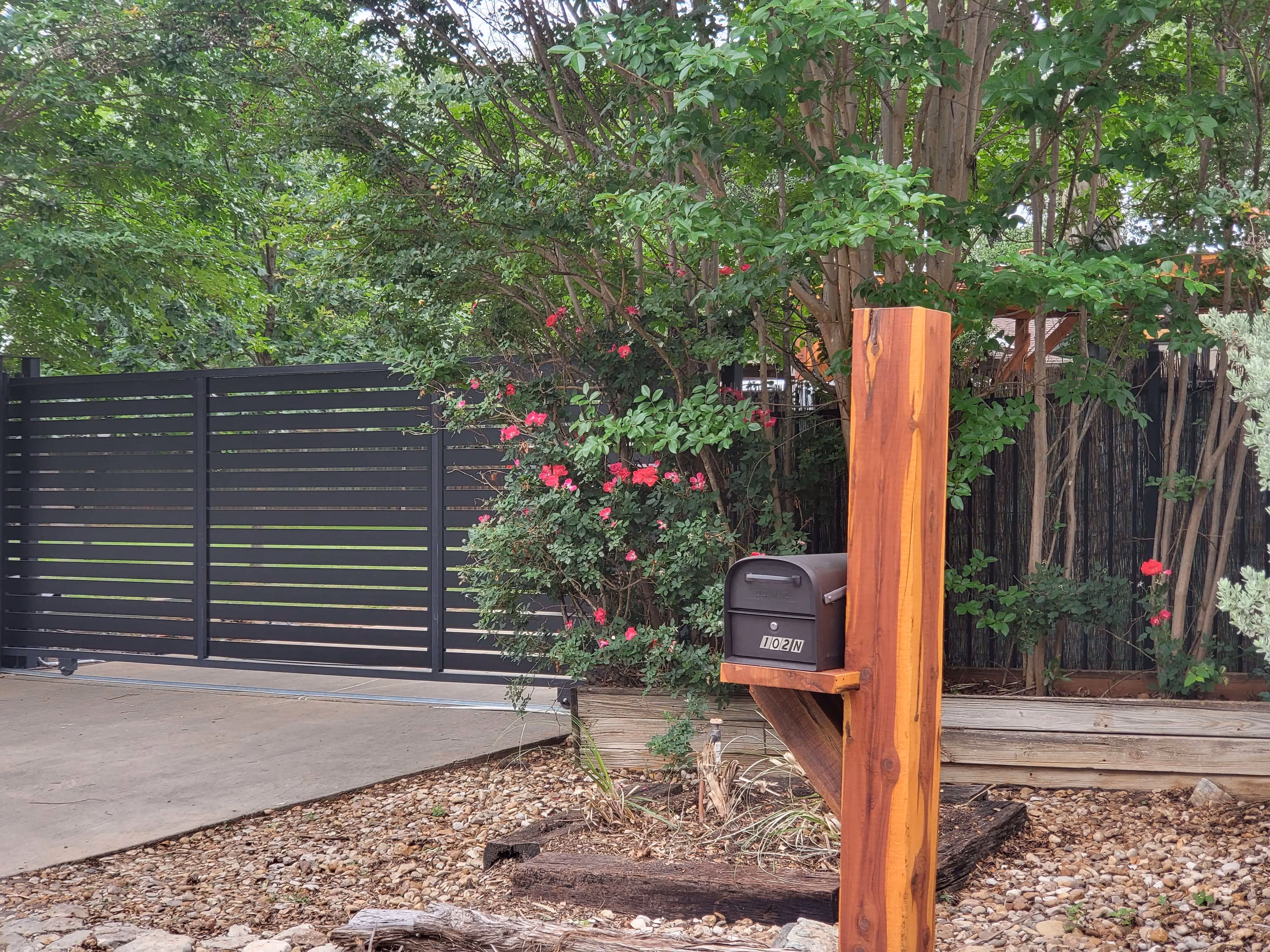Black aluminum rolling gate with horizontal slats beside a wooden mailbox post surrounded by greenery and pink flowers.