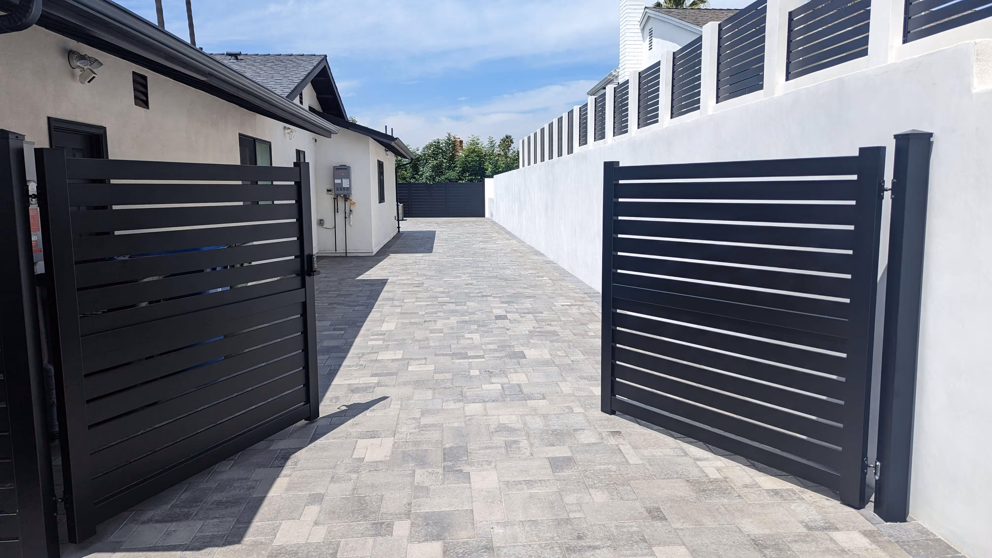 Open black horizontal aluminum double swing driveway gate on a tiled driveway beside a white house and white wall.