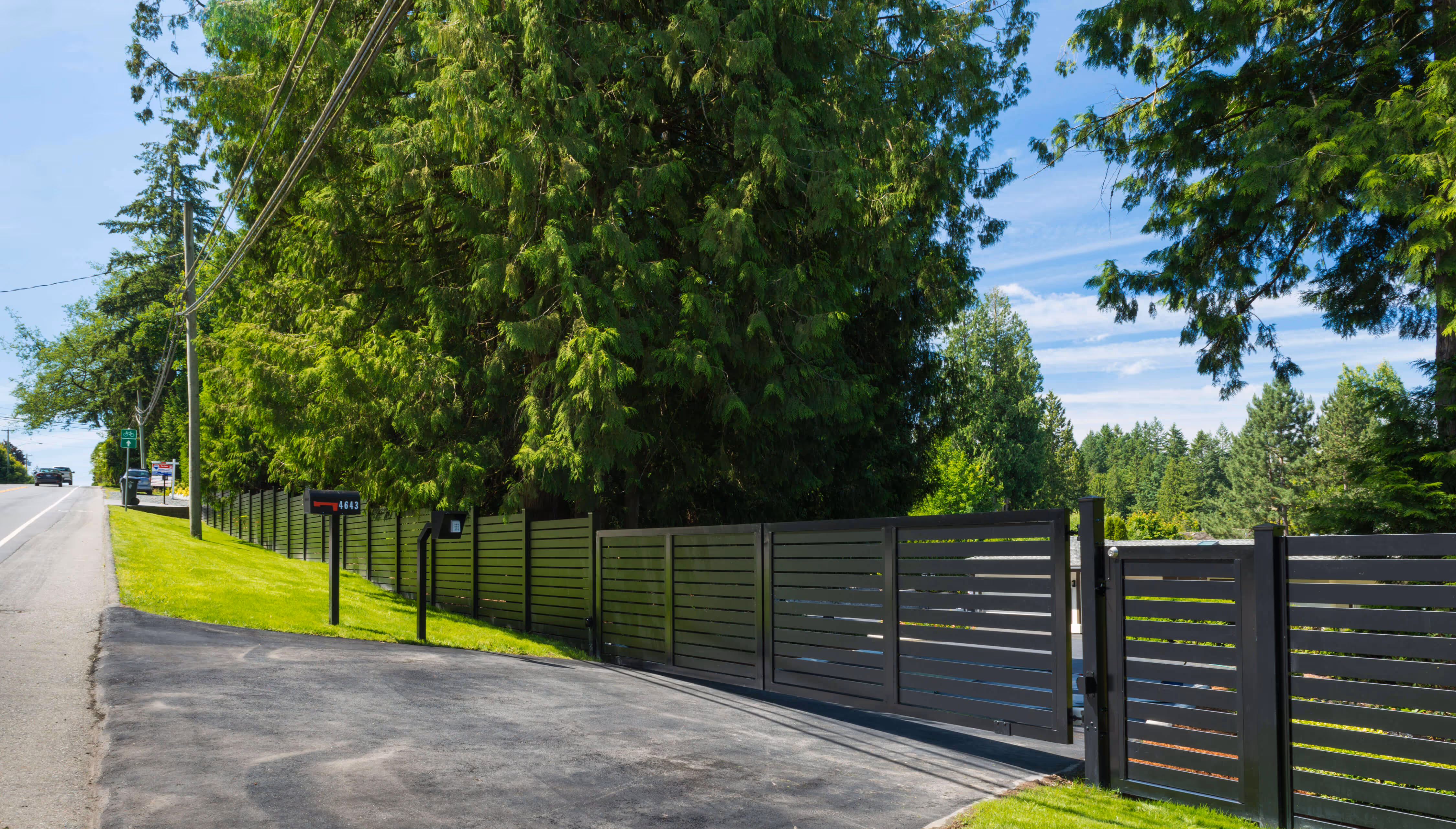 Modern black aluminum fence and gate along a sloped driveway beside green grass and tall trees with blue sky.