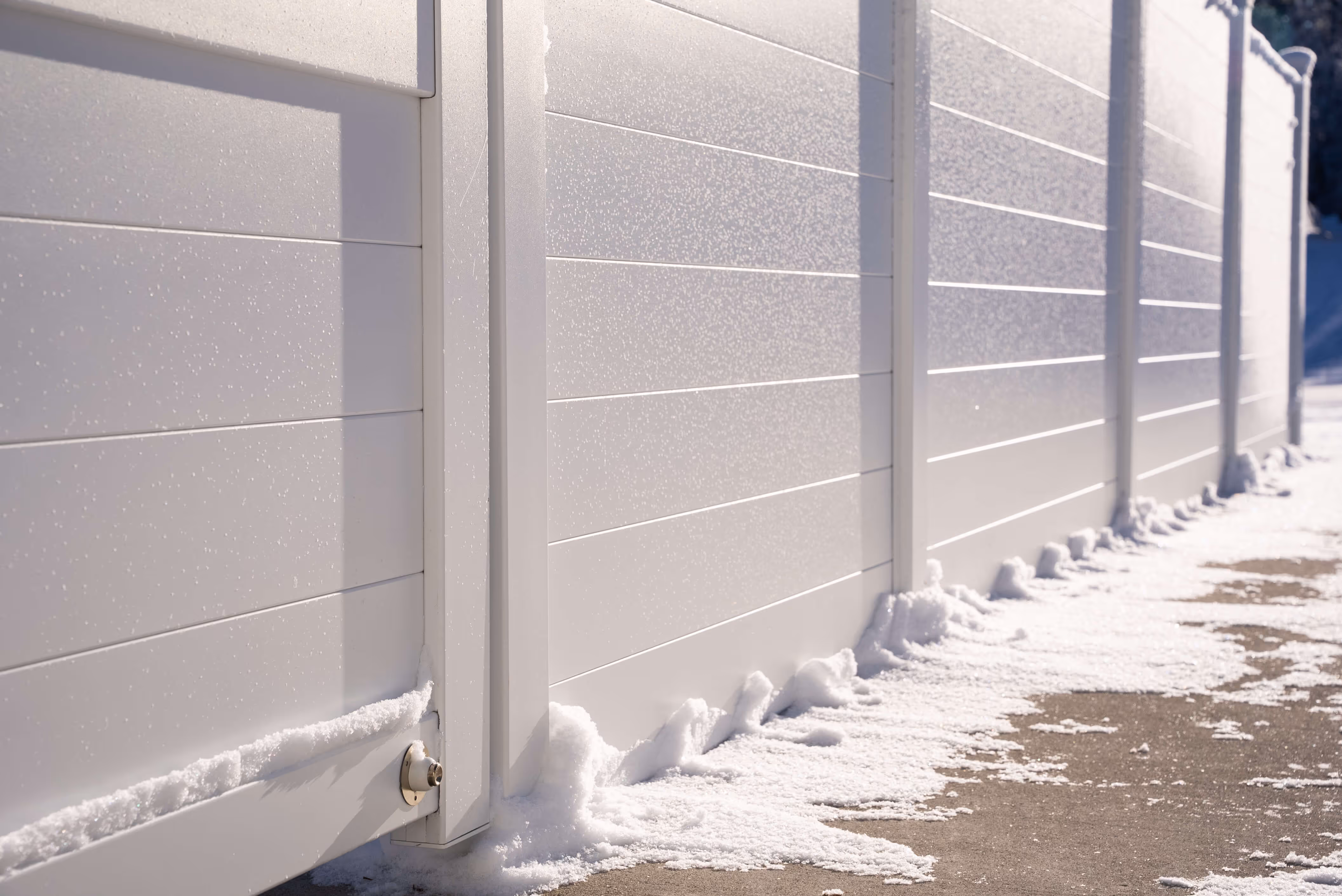 White horizontal panel fence with snow along its base on a sunny day.