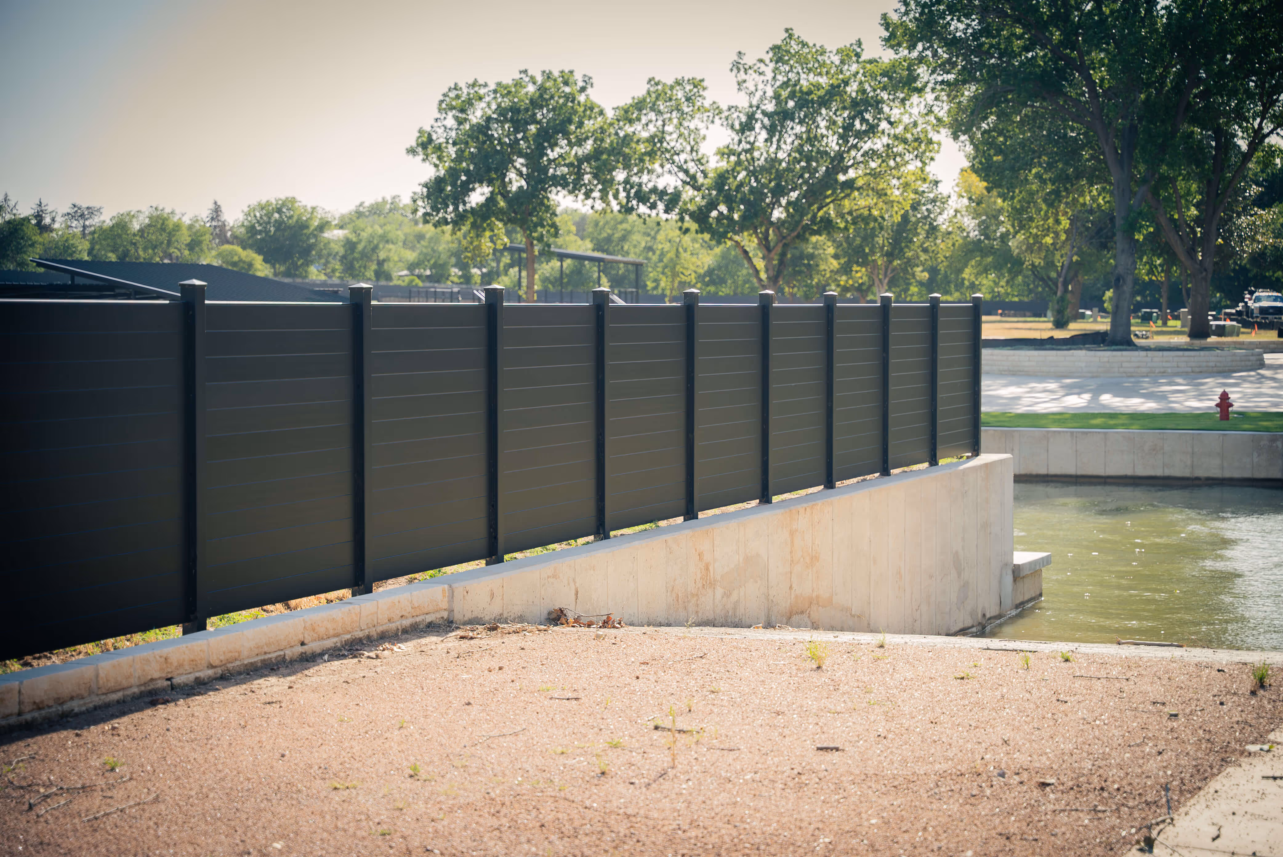 Black aluminum privacy fence installed along a concrete waterfront edge with trees and grass in the background.