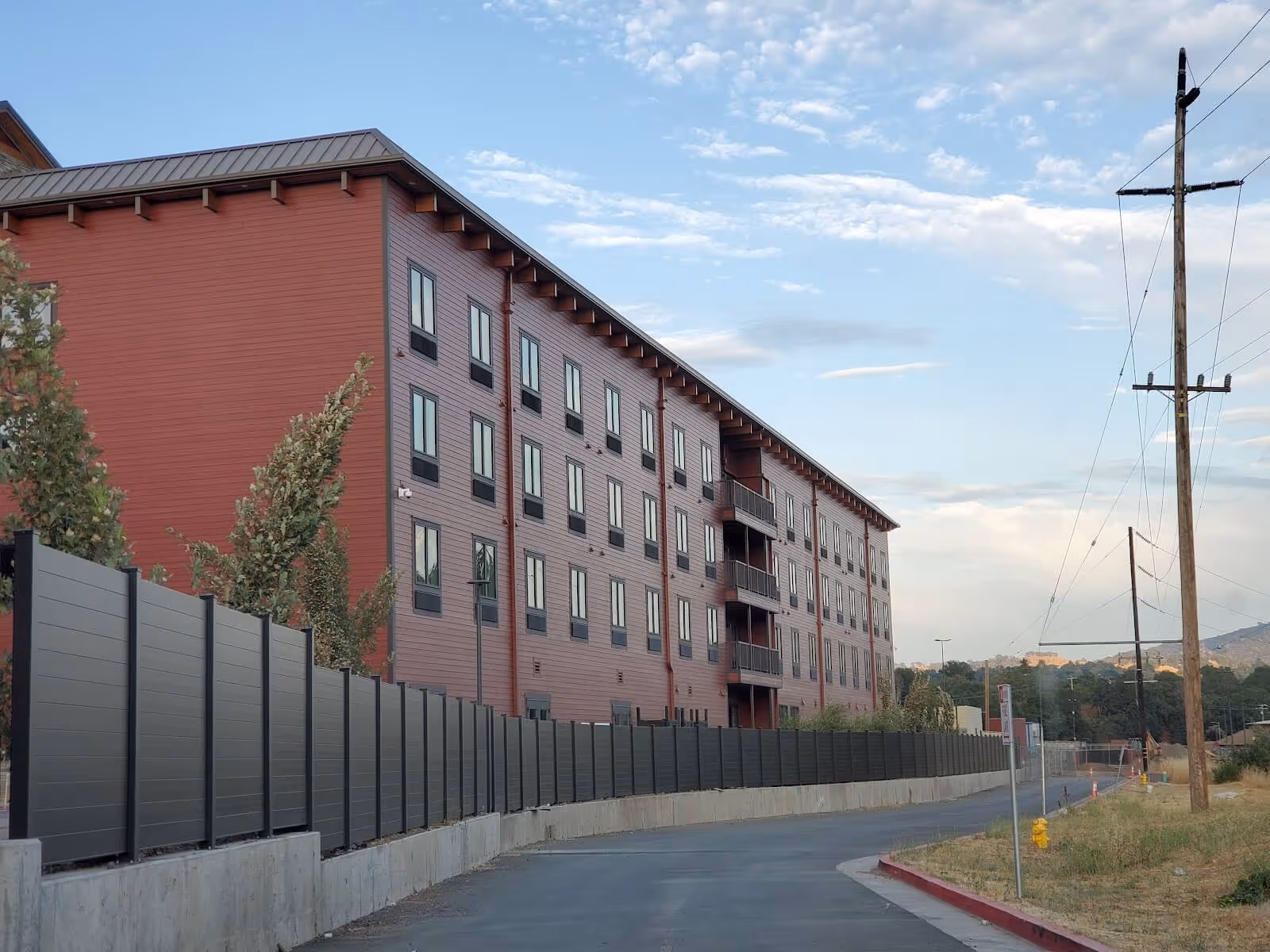 Modern red multi-story apartment building behind a long black aluminum fence along a curved street under a partly cloudy sky.