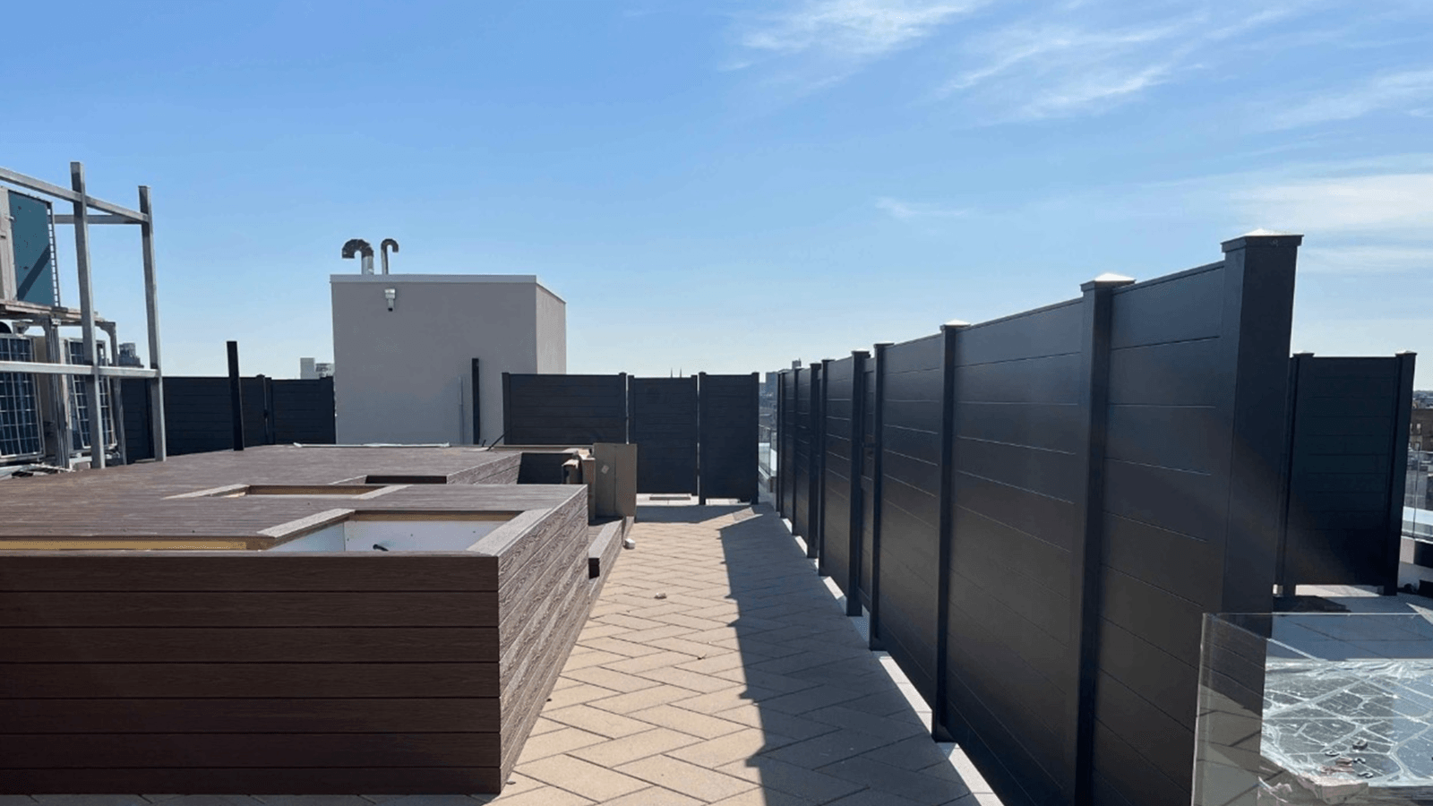 Rooftop area with light brown herringbone tile flooring, surrounded by tall black privacy fences and brown wooden planter boxes.