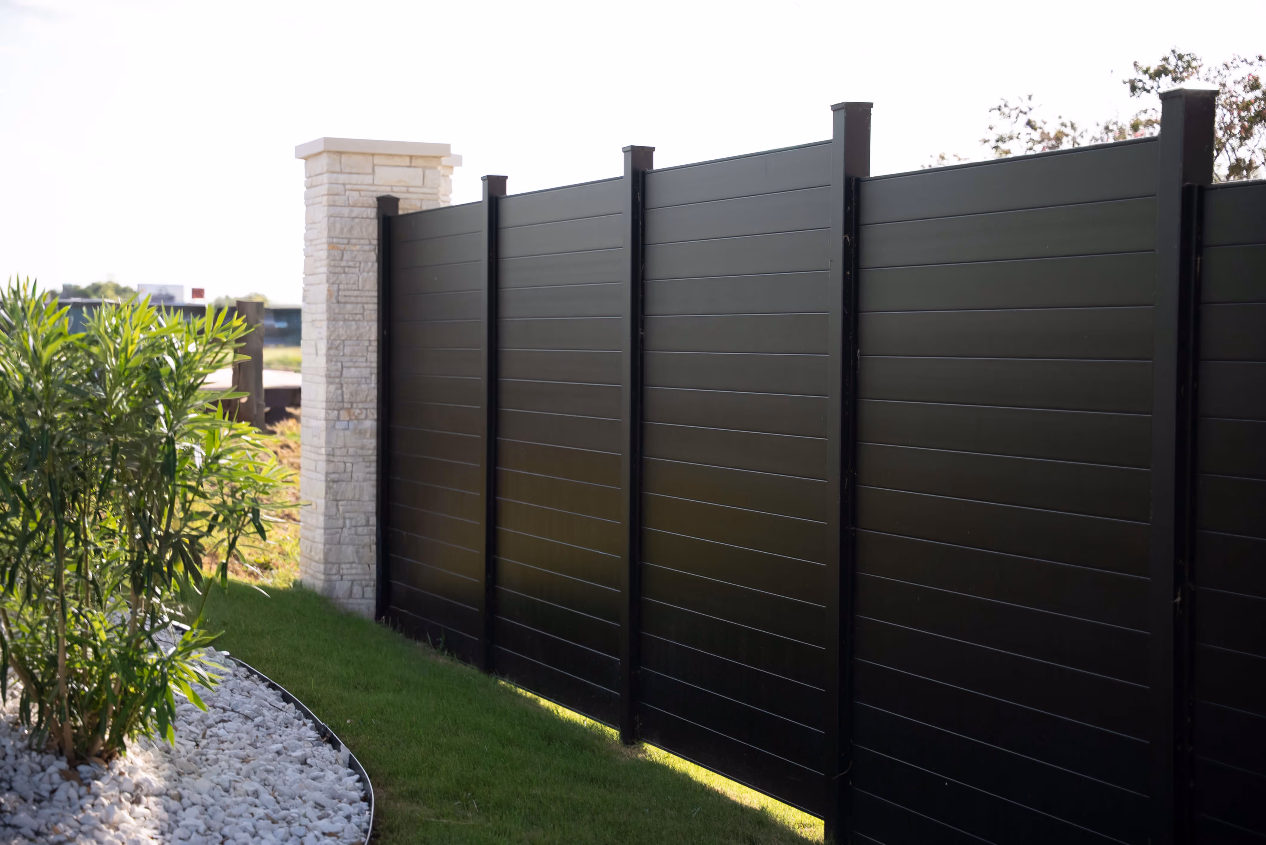 Modern black horizontal aluminum privacy fence next to a stone pillar and garden with green plants and white rocks.