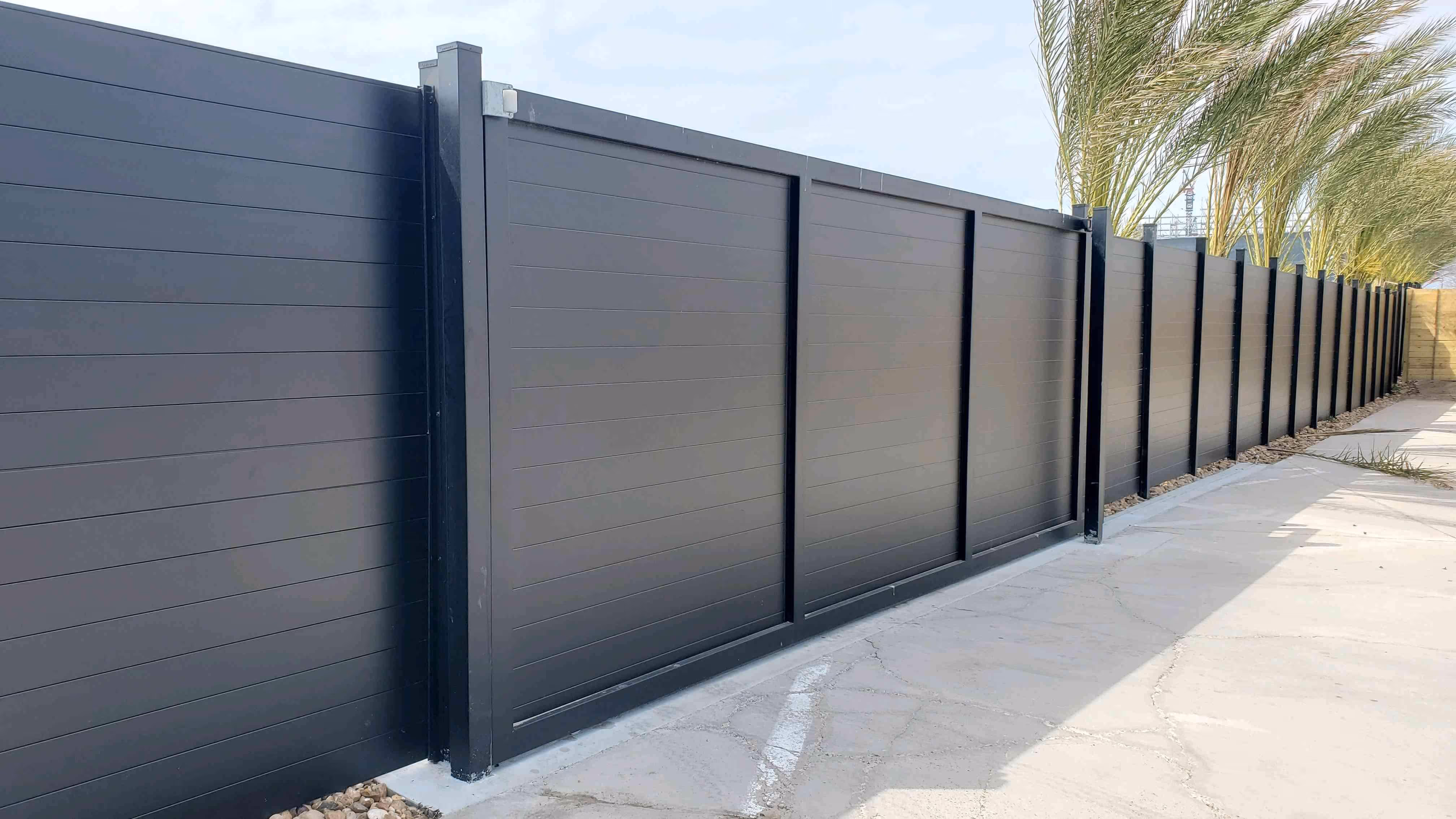 Modern black aluminum privacy fence with horizontal panels along a paved driveway and landscaped edge.