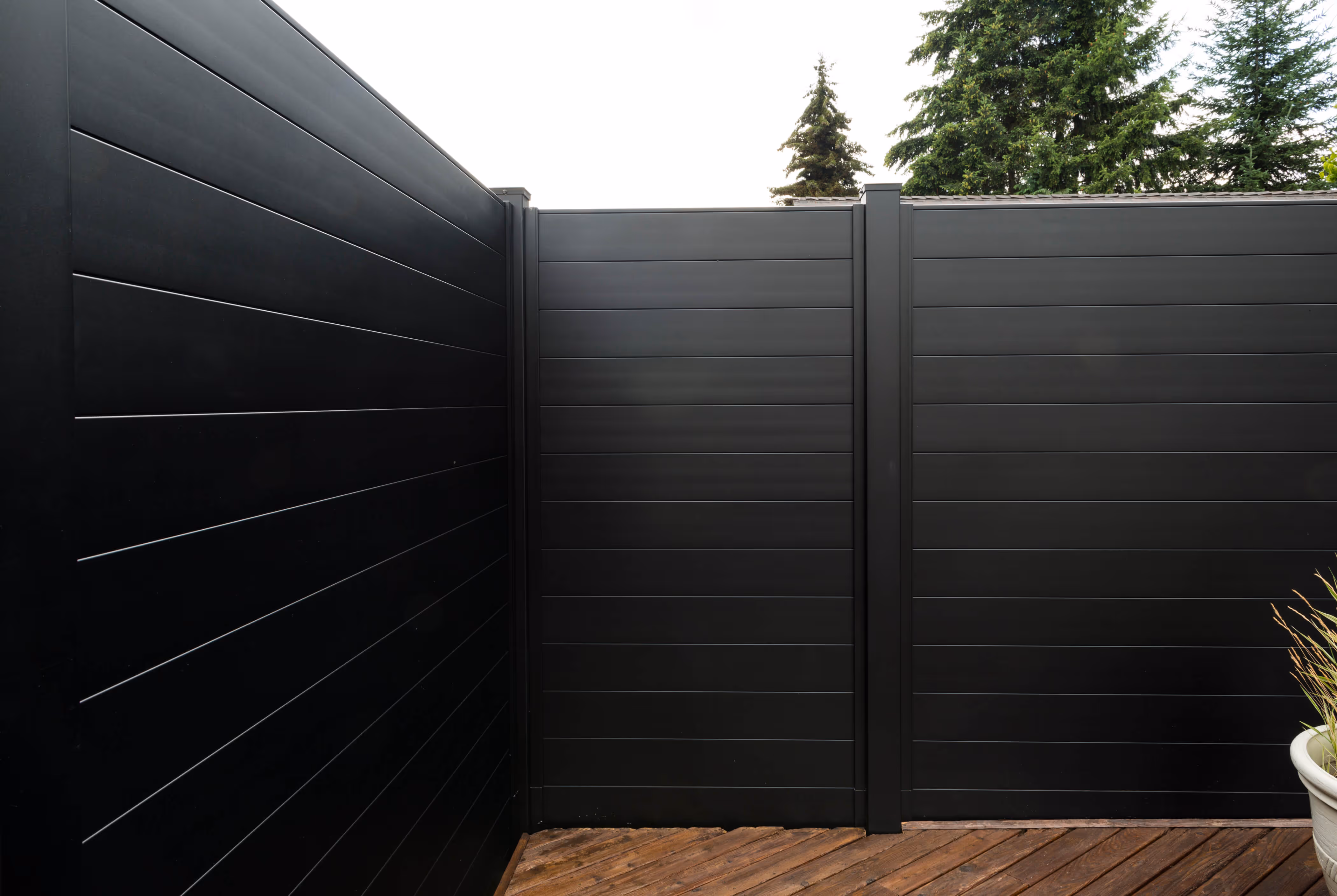 Modern black aluminum privacy fence with horizontal panels and dark wood deck, outdoor setting with trees in background.