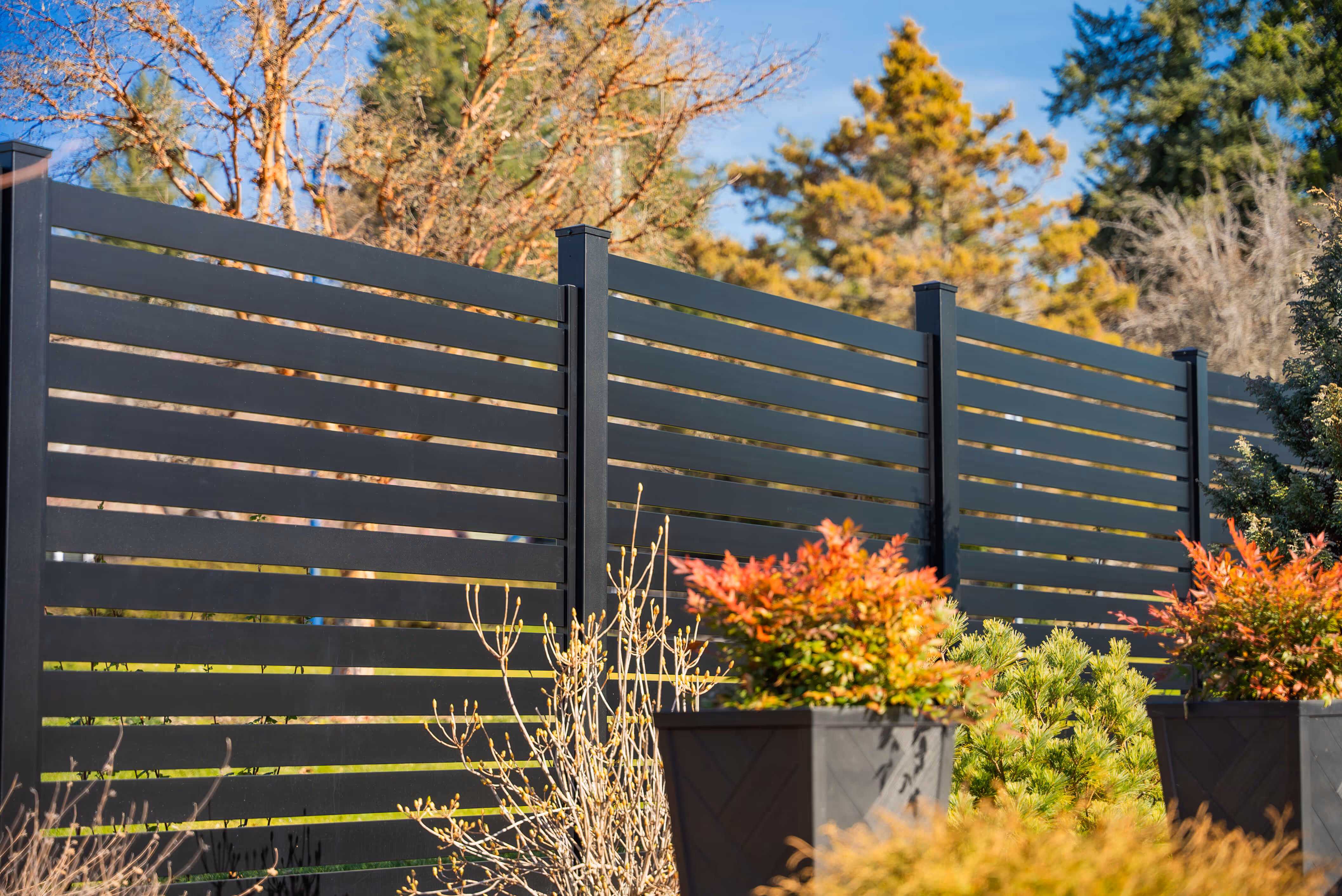 Black horizontal slat aluminum privacy fence with plants and shrubs in front under a clear blue sky.