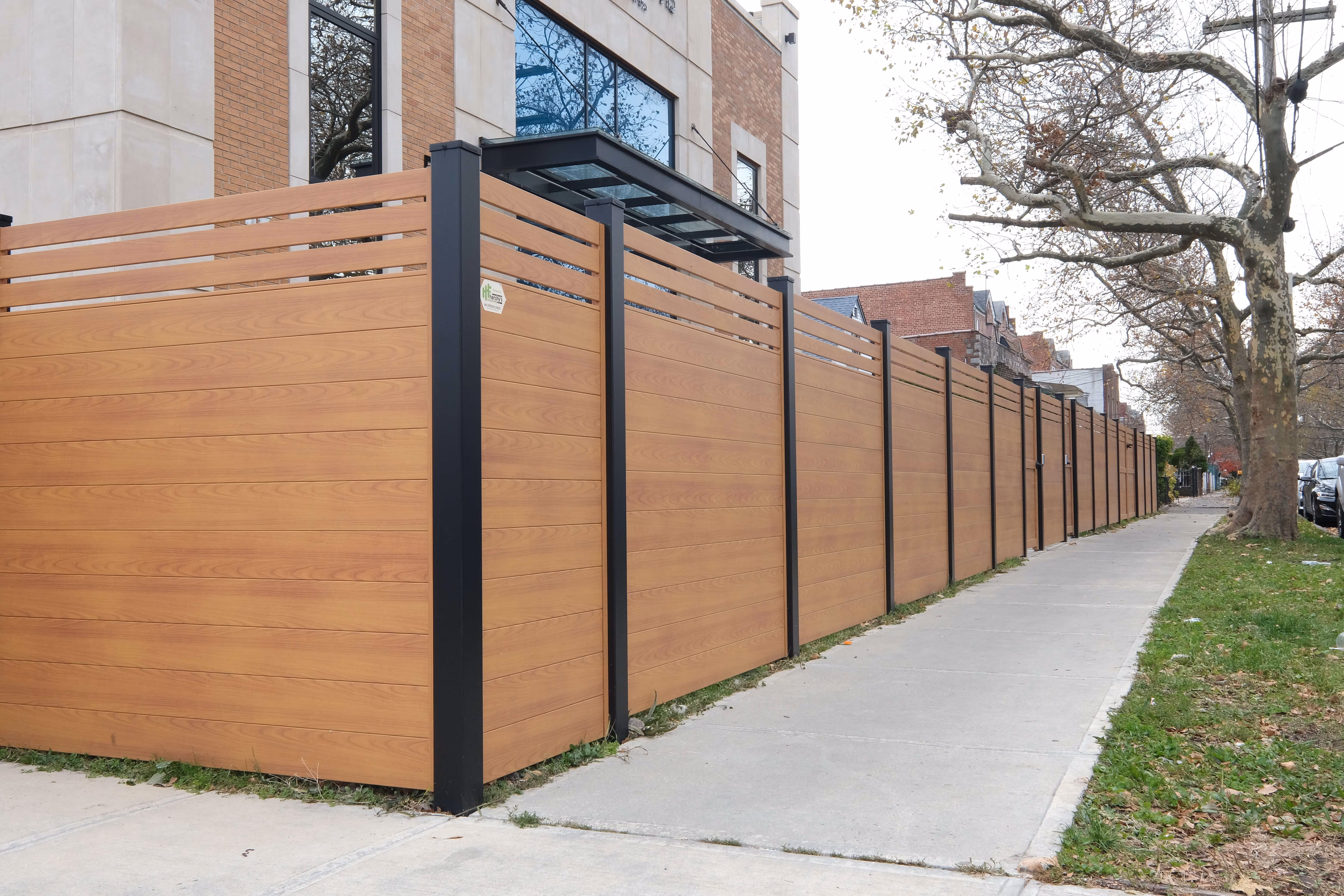 Long horizontal wood grain privacy fence with black metal posts alongside a sidewalk and trees.