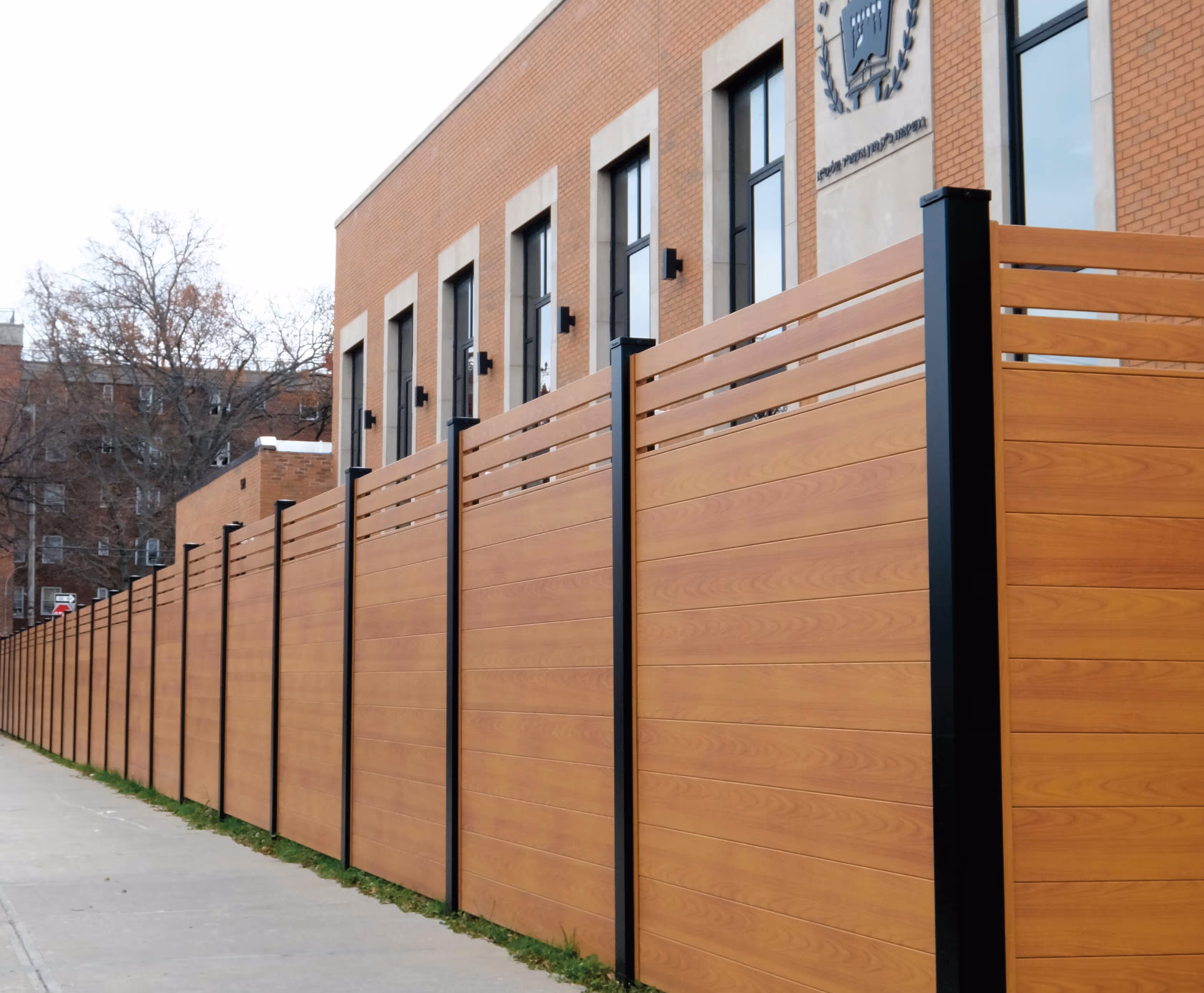 Long privacy fence with horizontal wood-grain panels and black vertical supports next to a brick building with large windows.