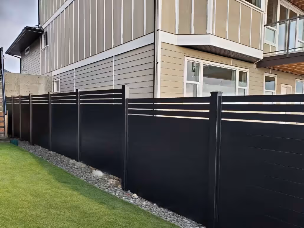 Black horizontal aluminum privacy fence with sleek design installed along a modern house with beige siding and a green lawn.