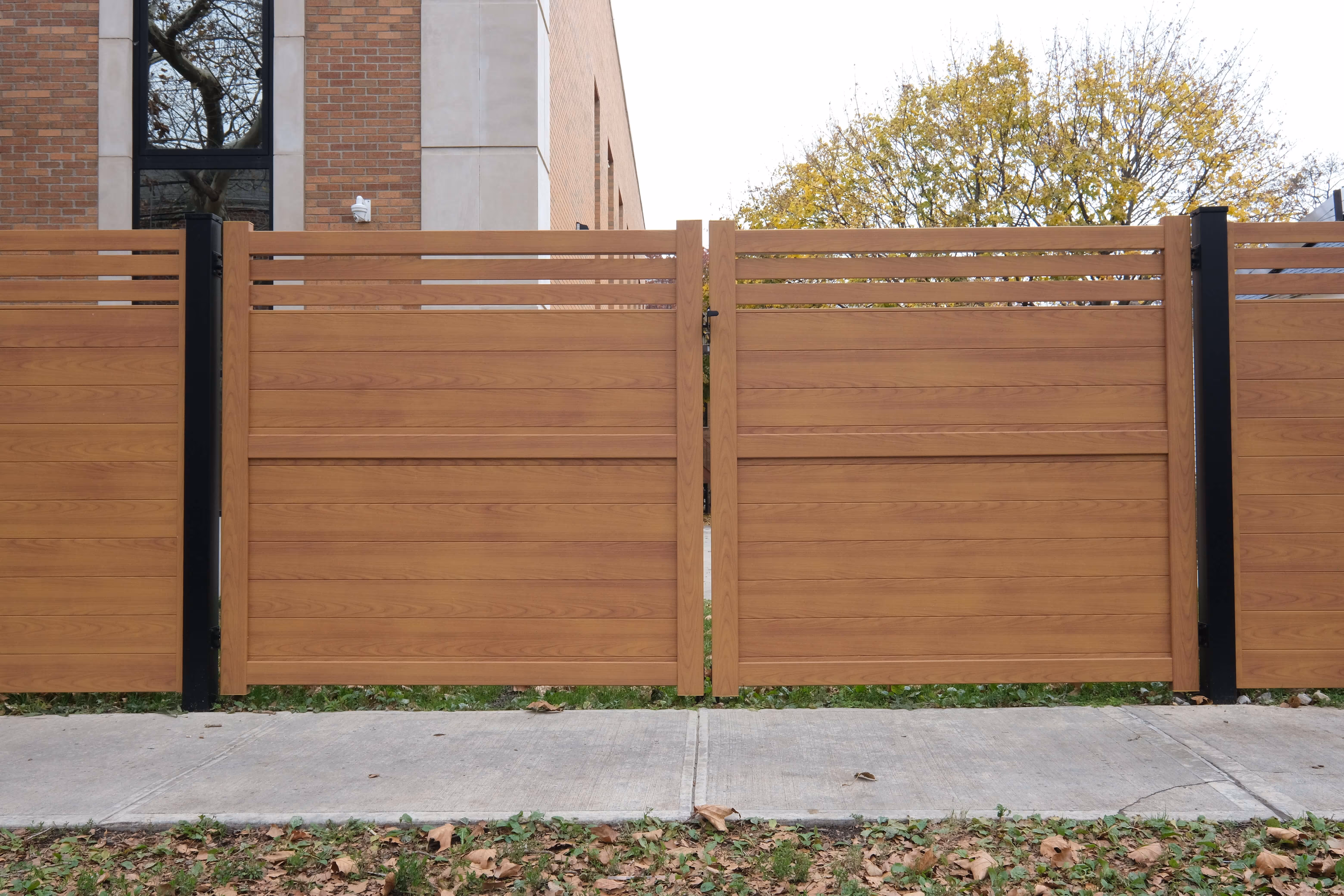 Modern horizontal aluminum vehicle gate with wood grain finish set between black posts in front of a brick building and trees.