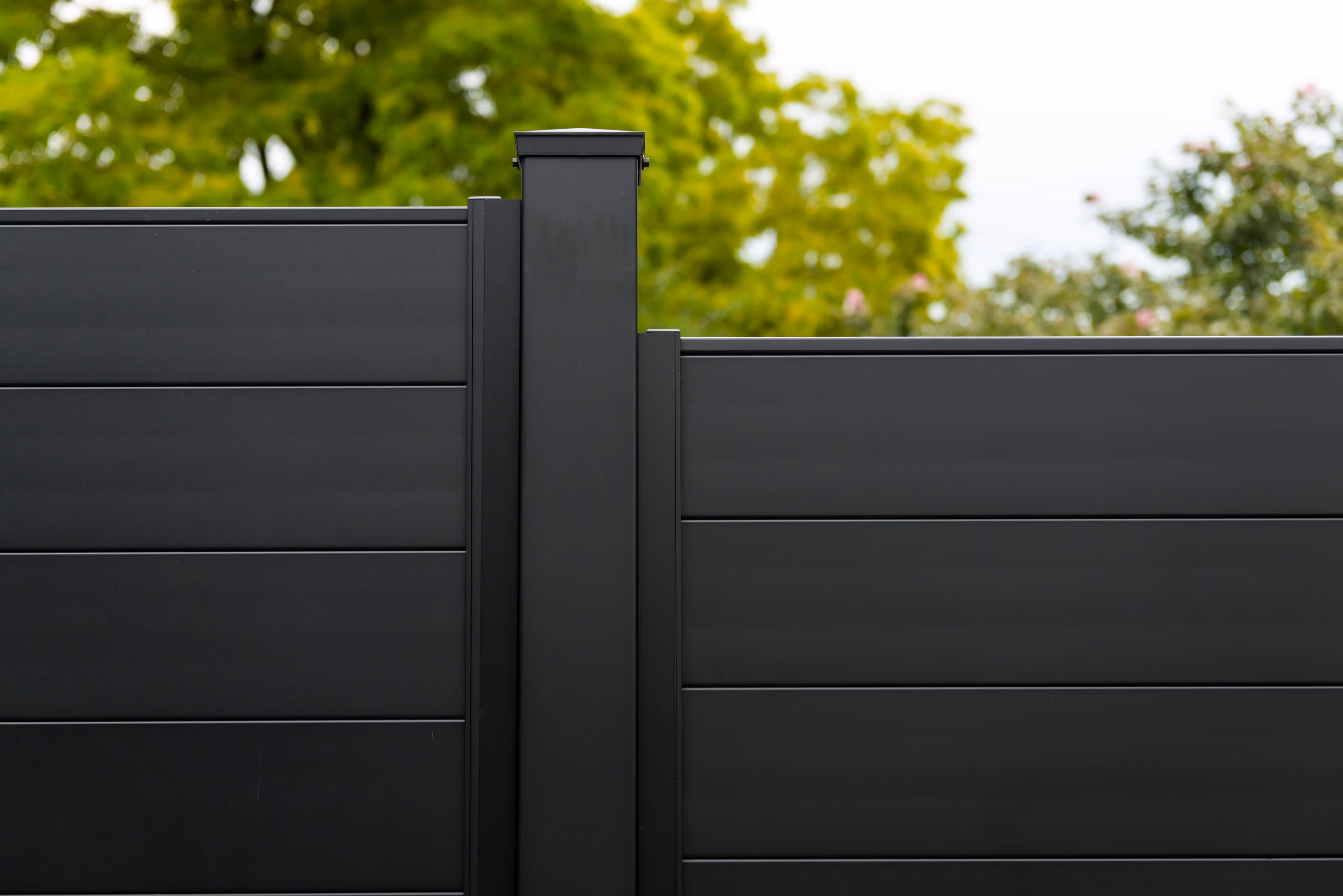 Close-up of a sleek black aluminum fence panel with horizontal slats and a vertical post, with green foliage in the background.