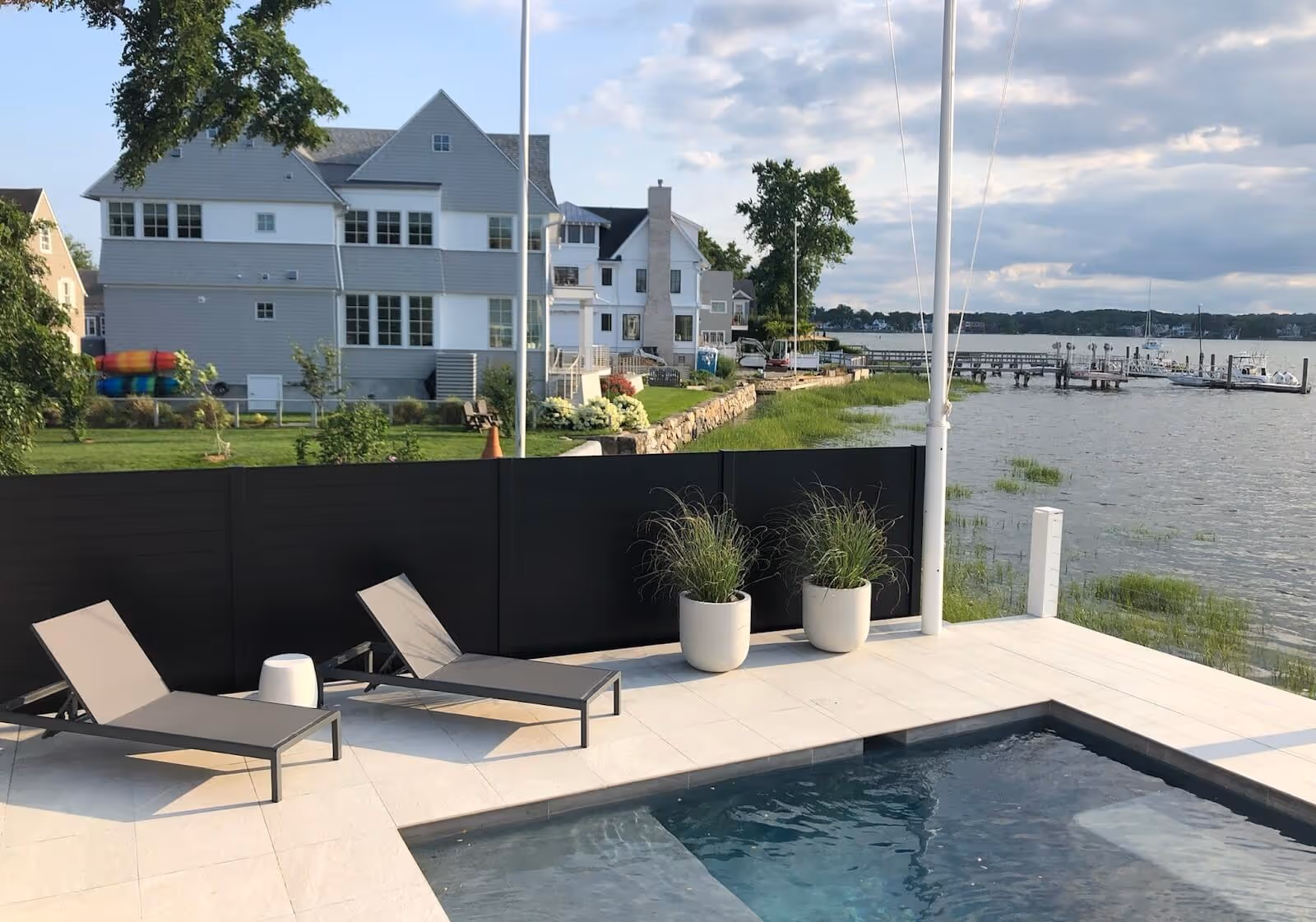 Modern poolside patio with two gray lounge chairs, black privacy fence, and a waterfront view with boats docked at a pier.