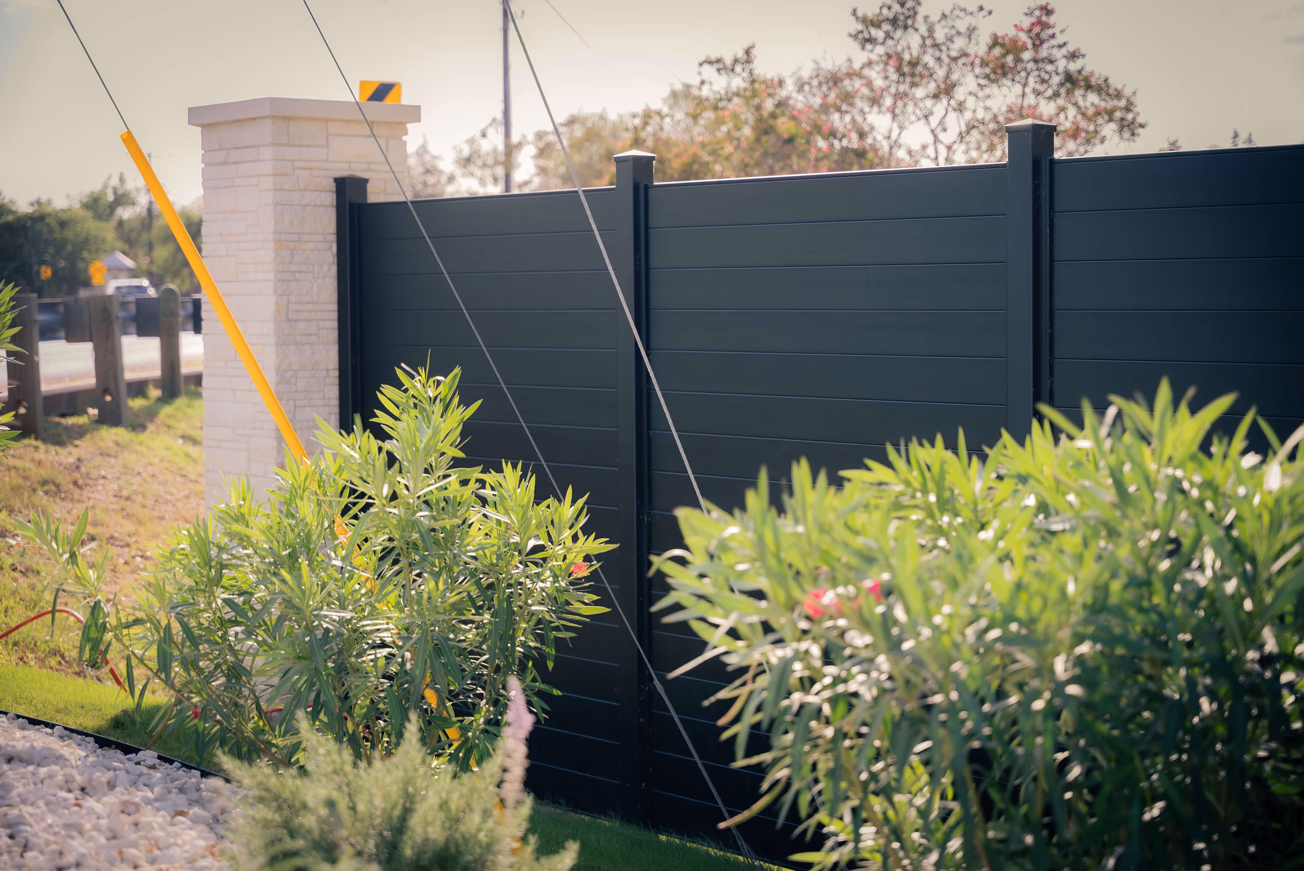Black aluminum privacy fence panels next to green shrubs and a stone pillar in an outdoor garden setting.