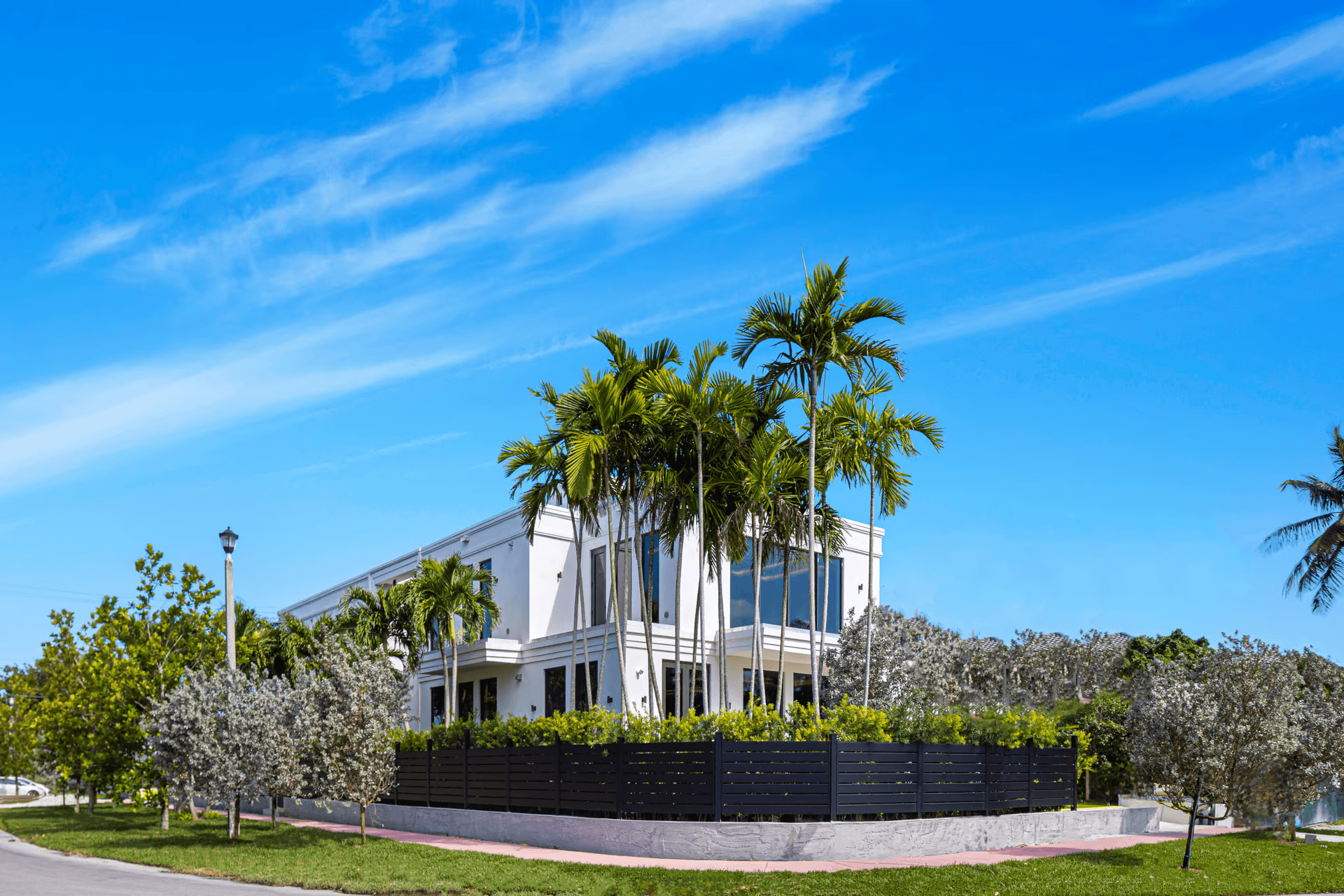 Modern white two-story house surrounded by palm trees and a black horizontal slat fence.