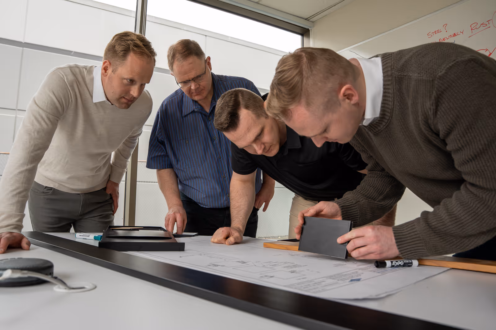Four men closely examining architectural blueprints and materials on a table in a bright office.