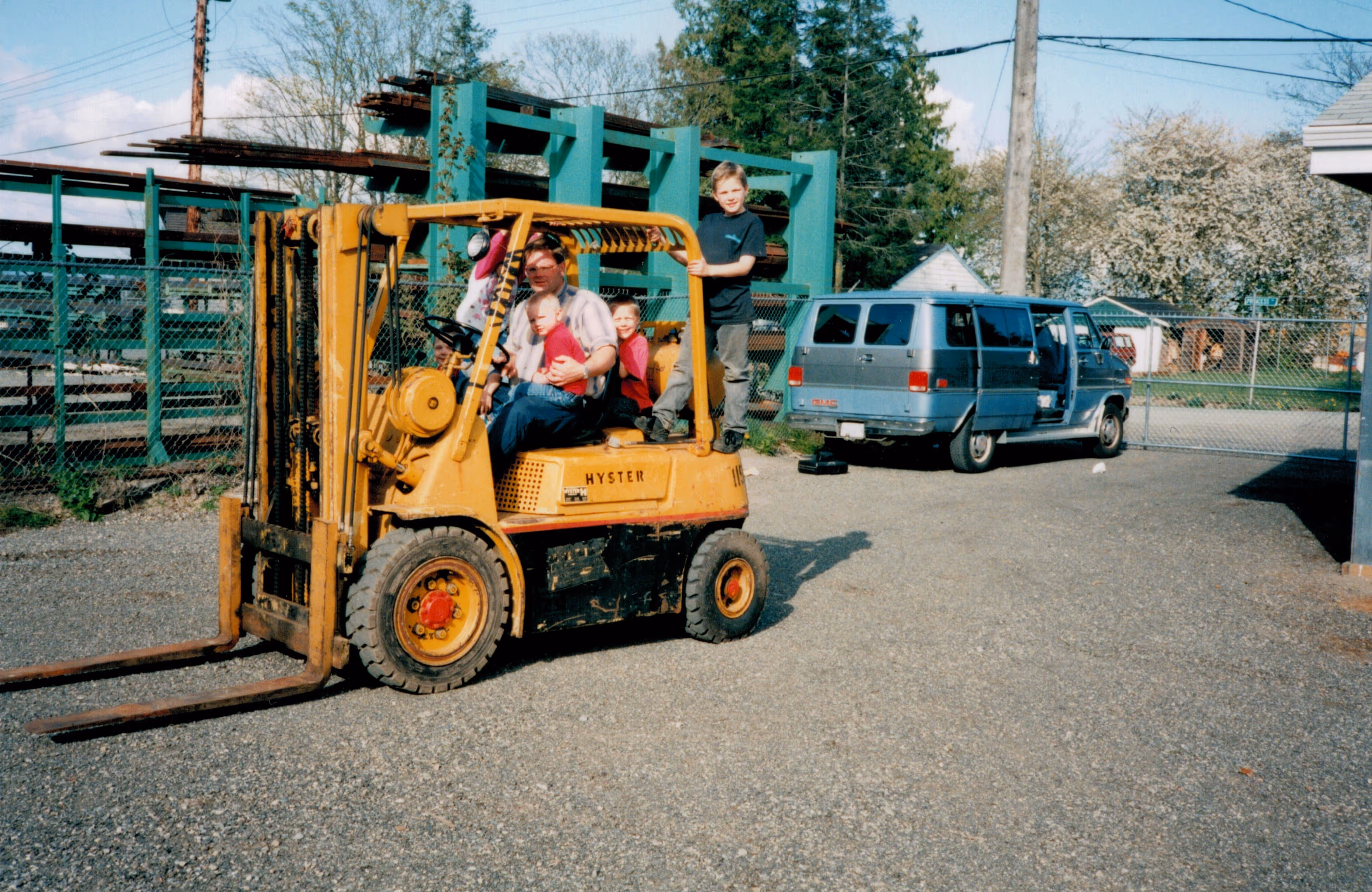 Man driving a yellow Hyster forklift with four children sitting and standing on it in an outdoor industrial area.