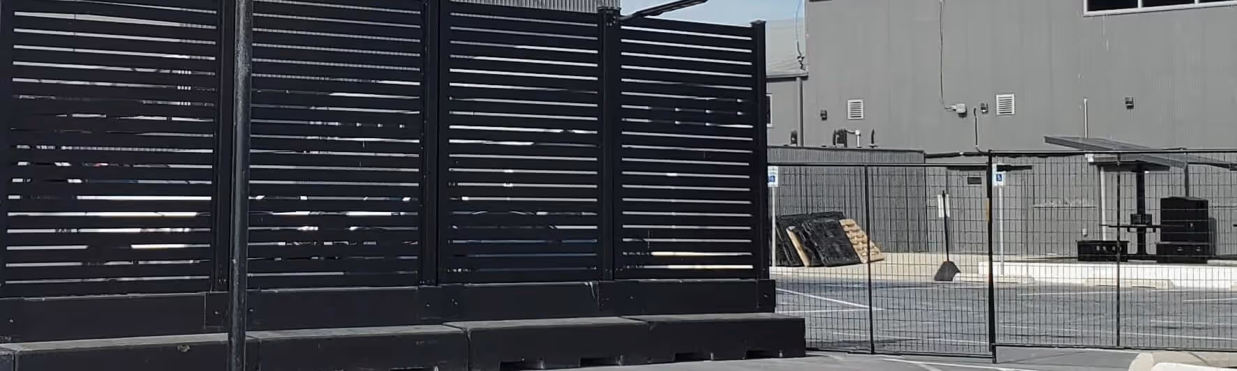 Black aluminum mobile fence panels with horizontal slats set on a concrete surface with a building and other fencing in the background.