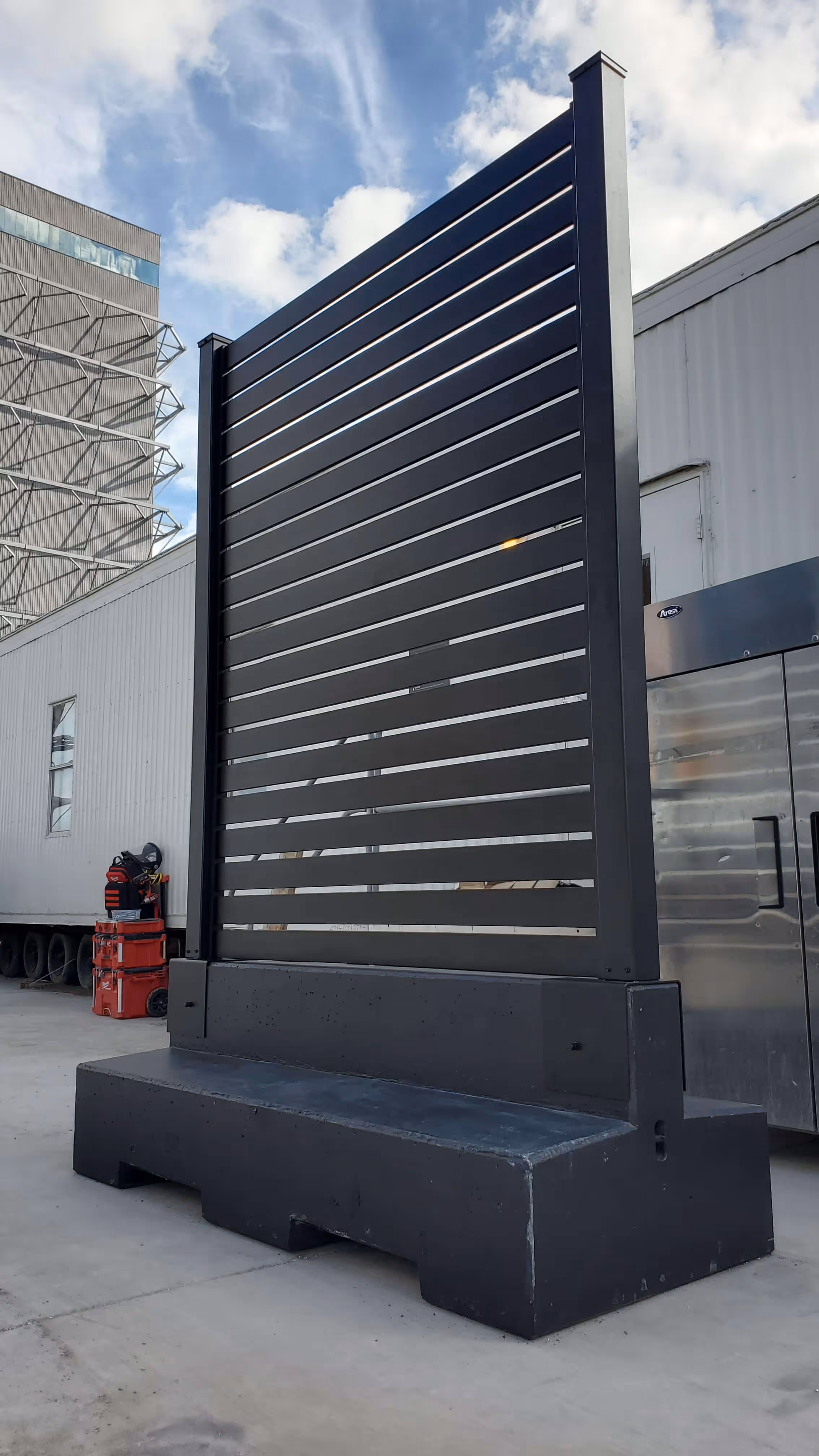 Black aluminum mobile semi-privacy fence panel mounted on a large concrete base outdoors.