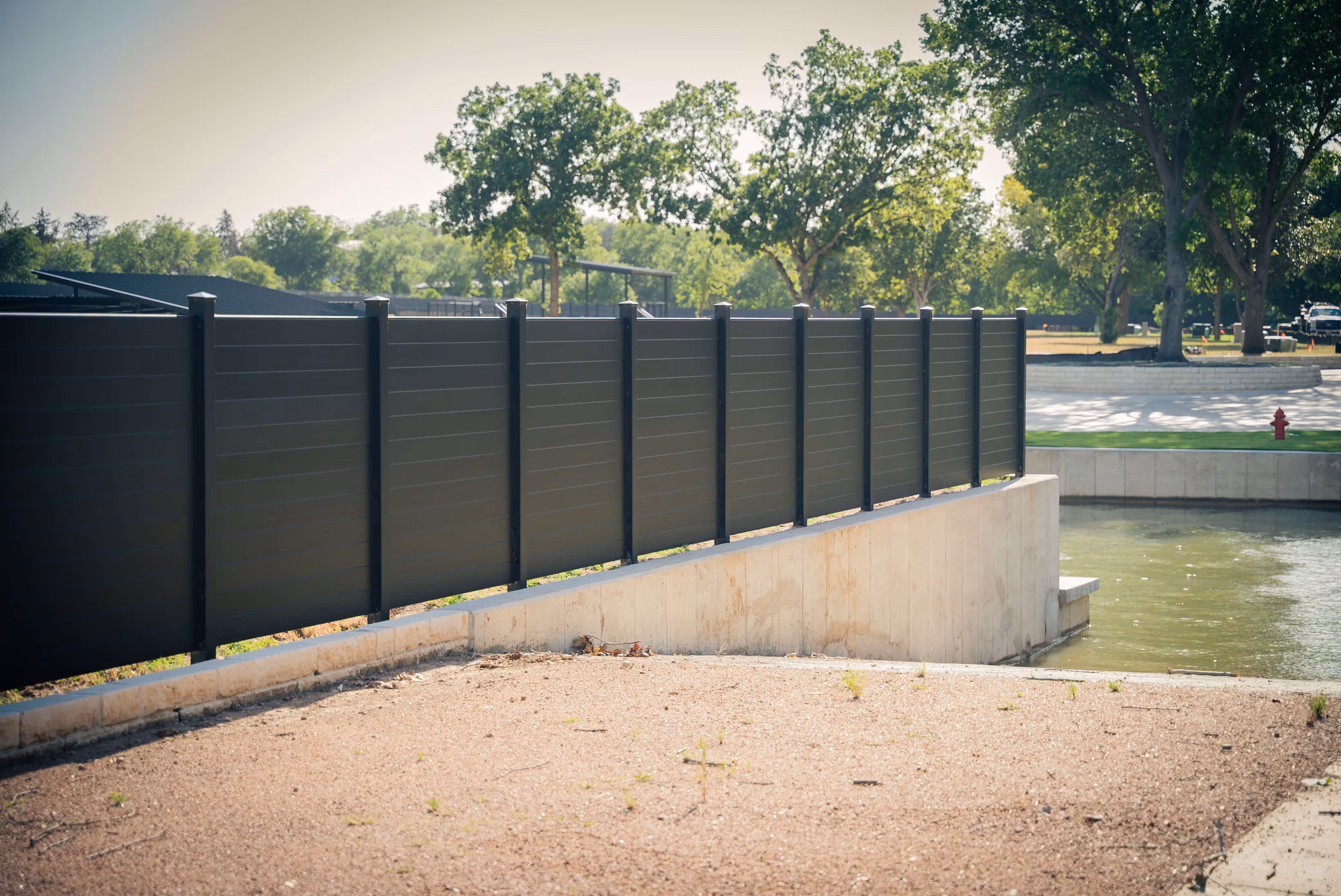 Black aluminum privacy fence installed on a concrete ledge beside a waterfront with green trees in the background.