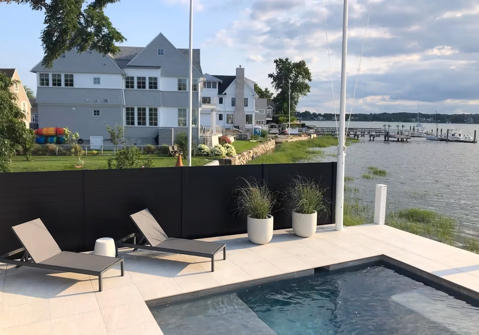 Modern outdoor poolside area with two lounge chairs, potted plants, black fence, overlooking waterfront houses and a dock.