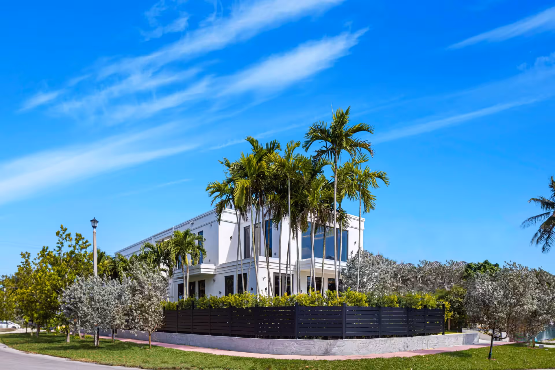 Modern two-story white house surrounded by palm trees and a horizontal black aluminum fence under a blue sky.