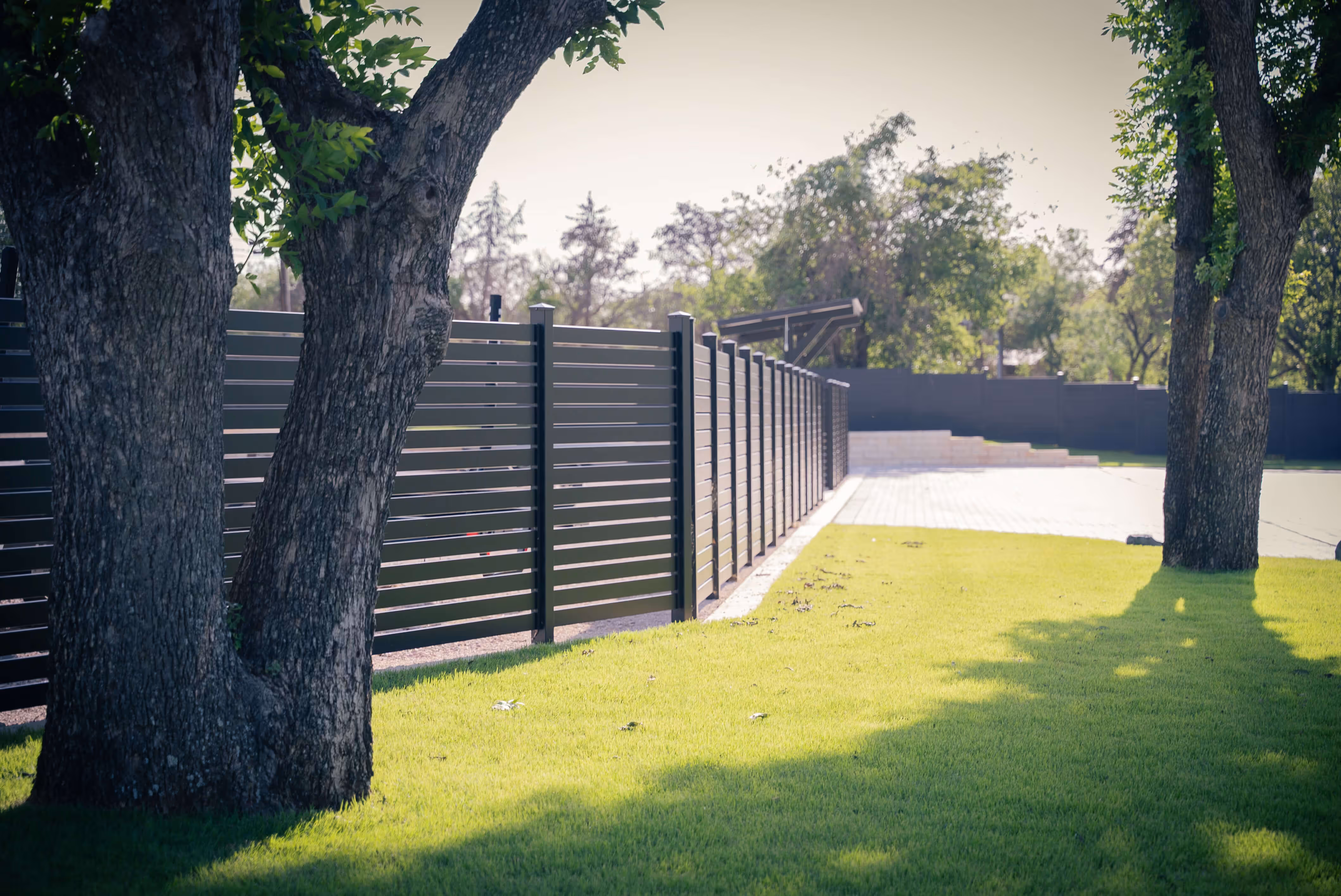 Black horizontal slat aluminum fence alongside a green lawn with two large trees in the foreground.