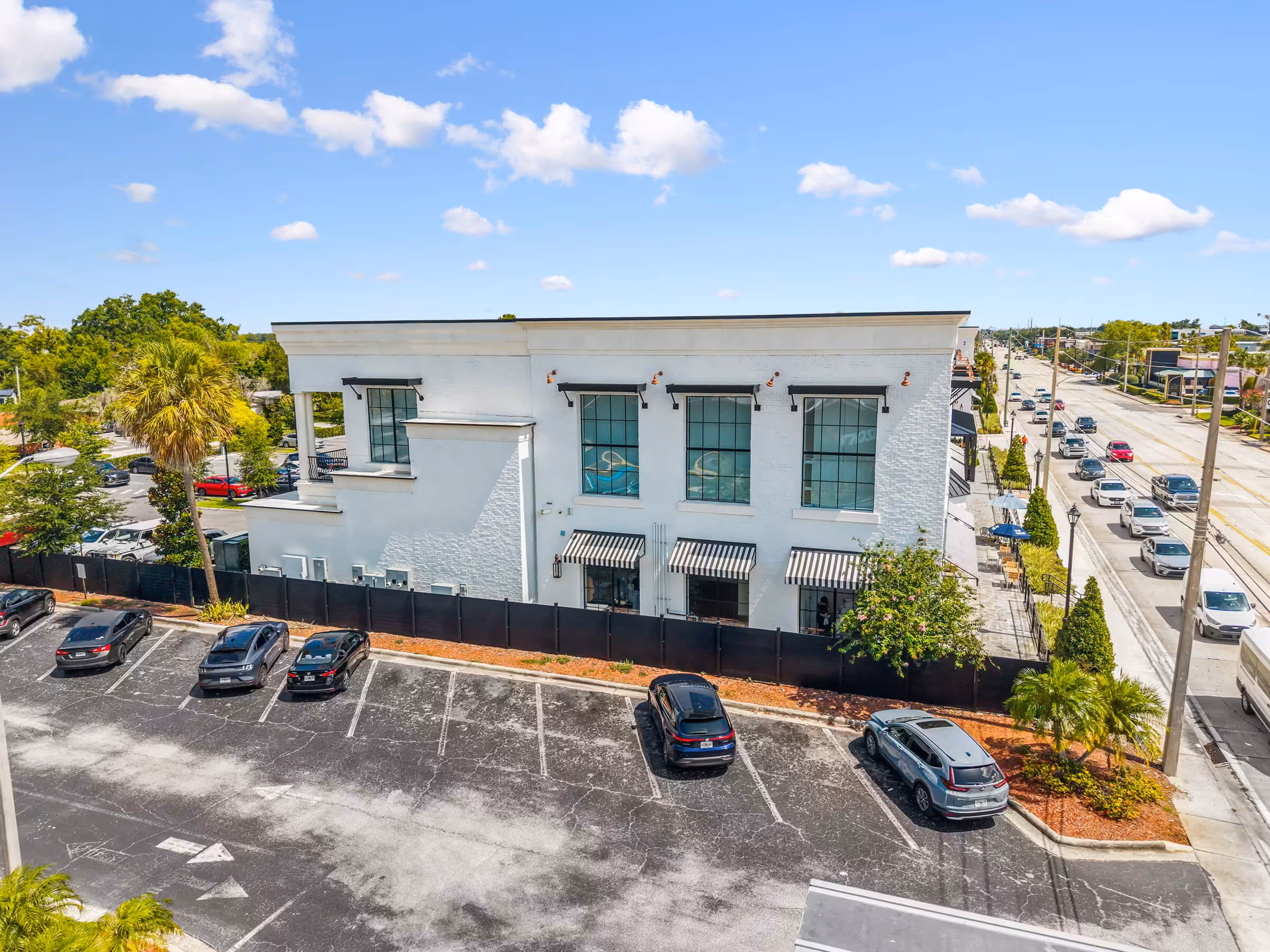 White two-story commercial building with large windows, black fence, and parked cars in front under a blue sky.