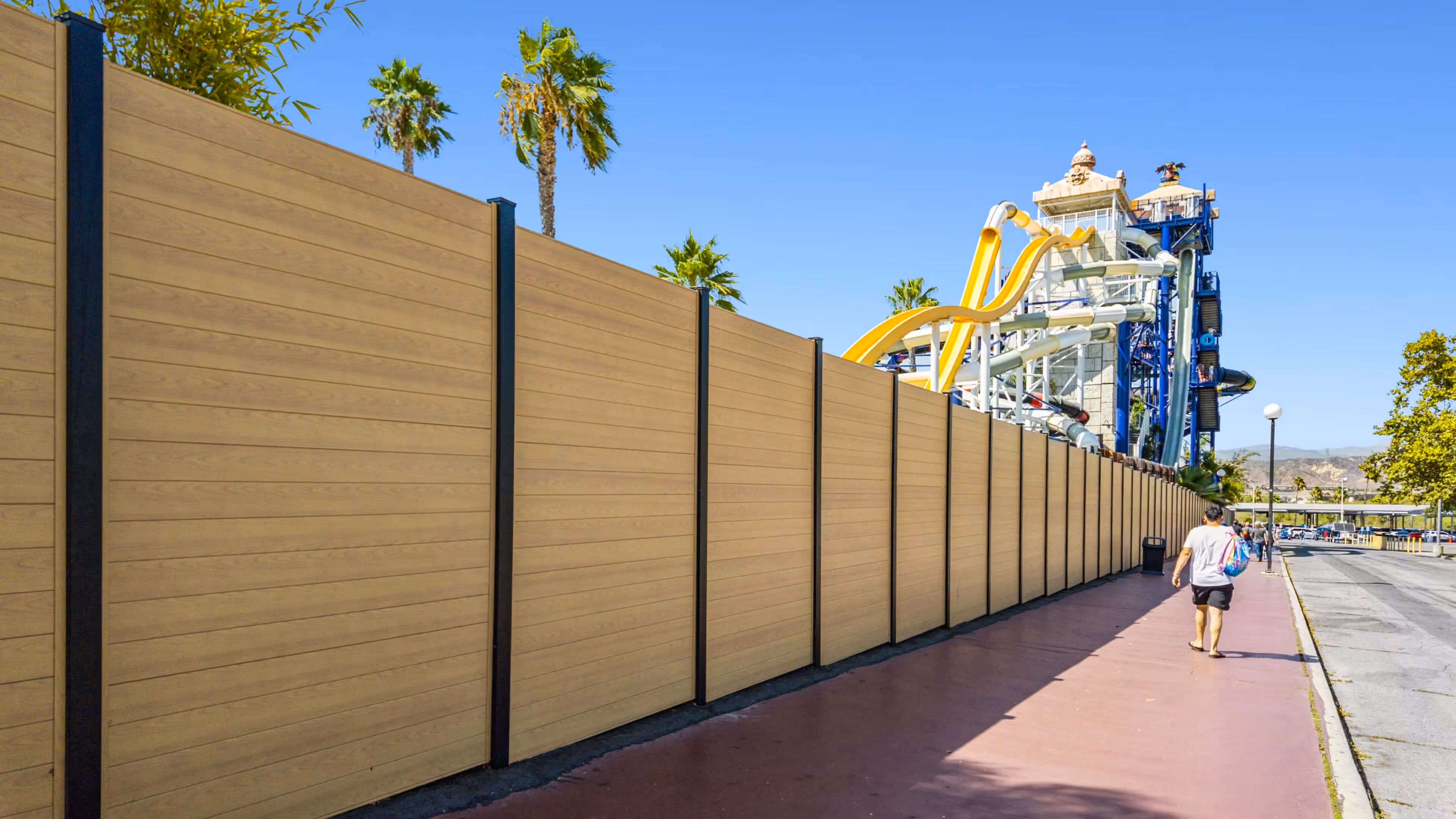 Long tan wind-resistant aluminum privacy fence alongside a sidewalk with a water slide structure and palm trees in the background under a clear blue sky.