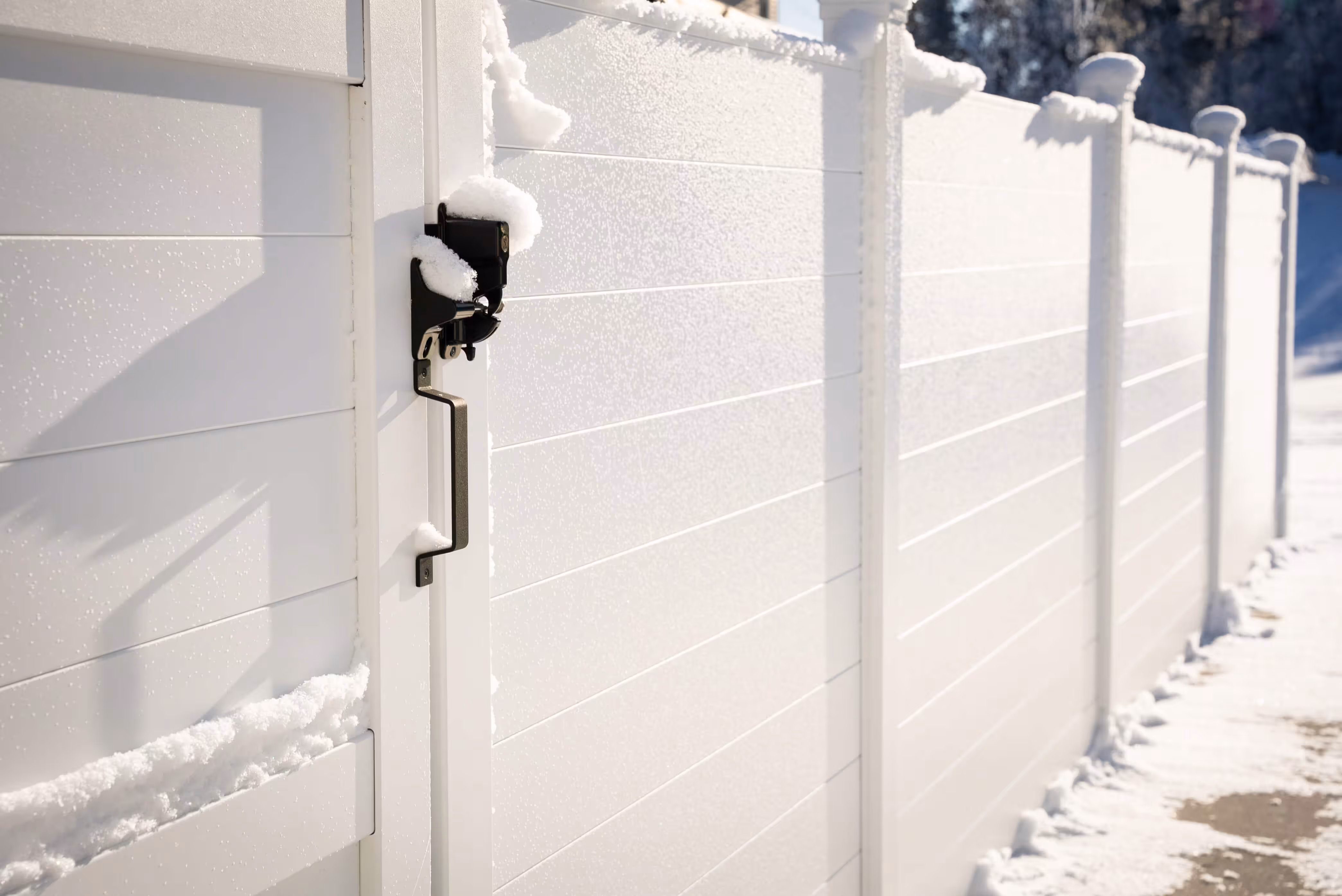 White aluminum privacy fence with a black metal handle and lock partially covered in snow on a sunny winter day.