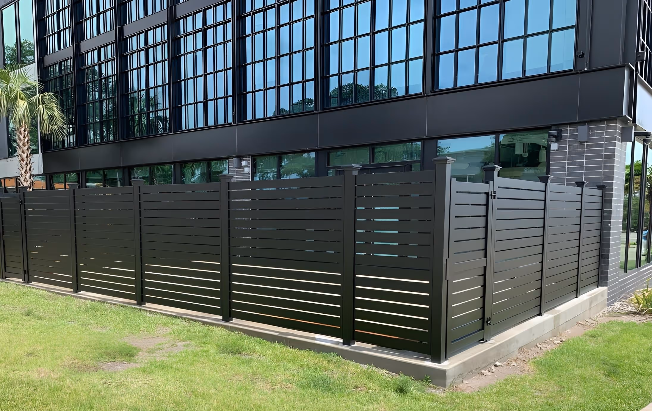 Modern black horizontal aluminum fence enclosing a corner of a commercial building with reflective glass windows.