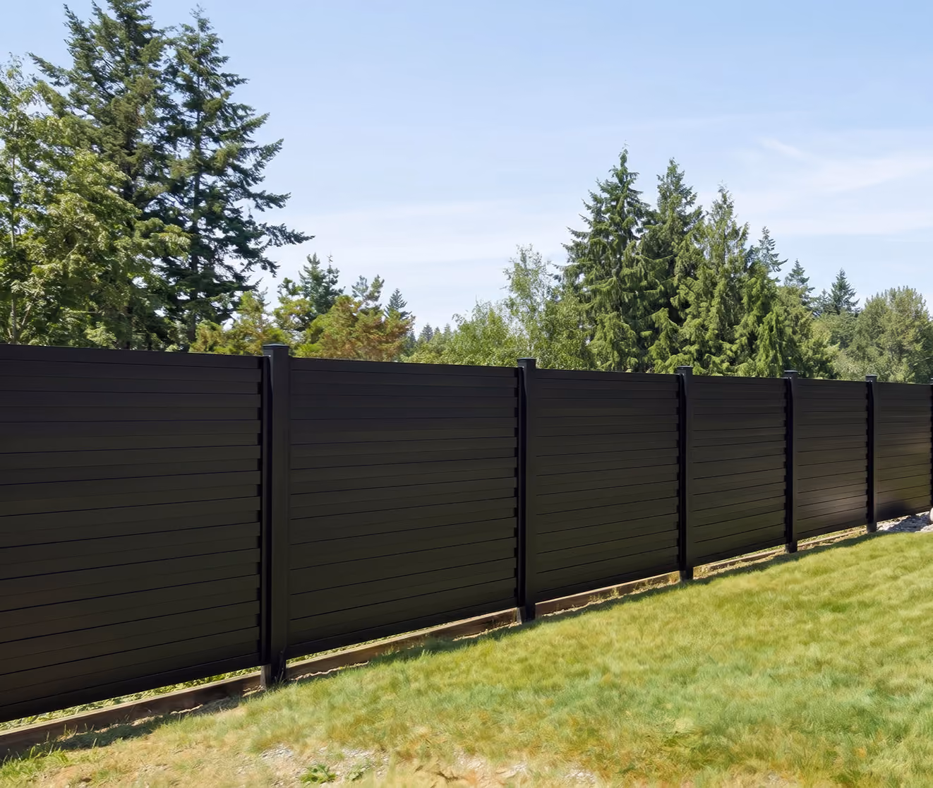 Black horizontal aluminum fence panels standing on green grass with trees and clear sky behind.