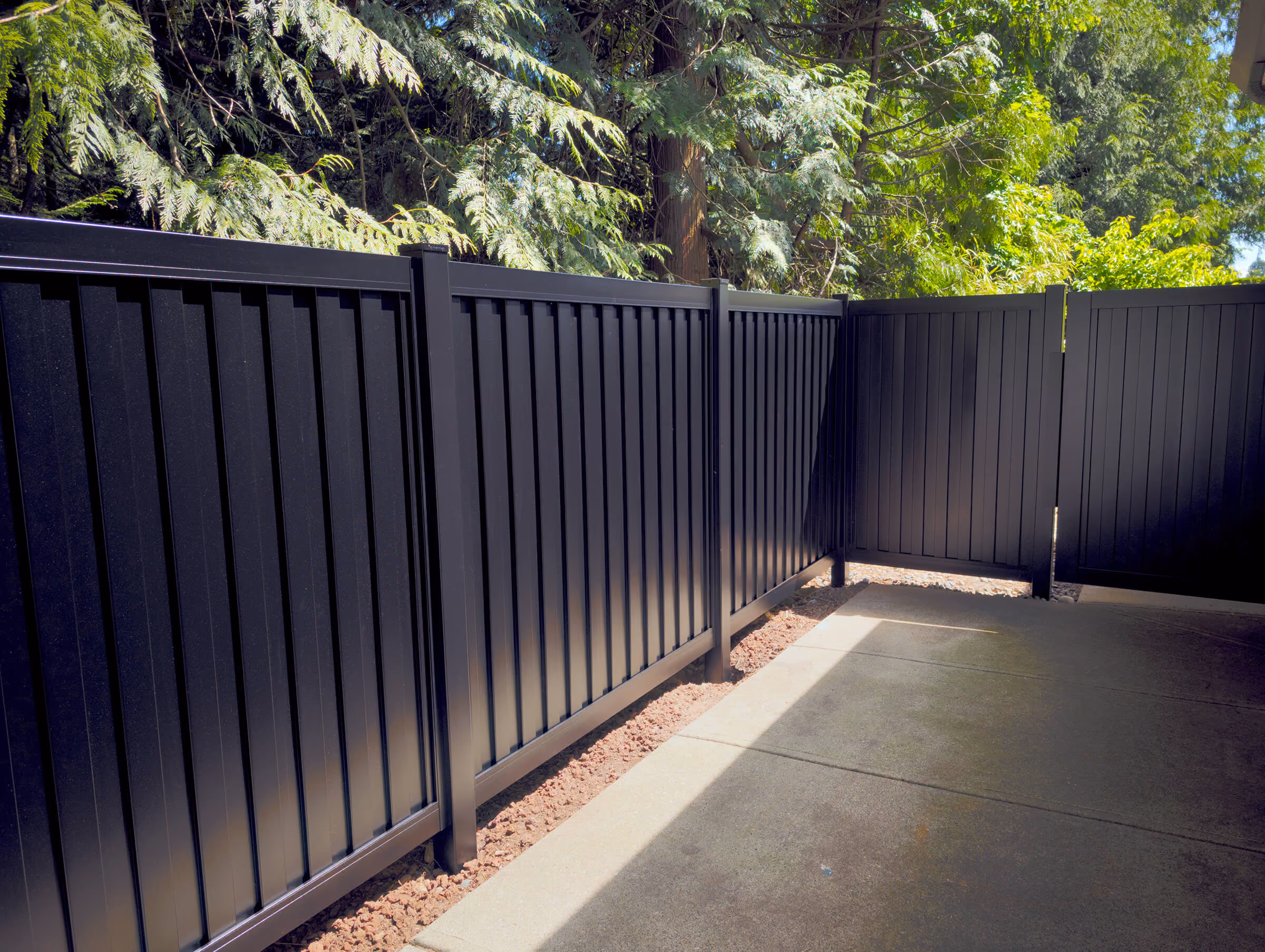 Black aluminum privacy fence and gate bordering a concrete patio with trees and greenery behind.