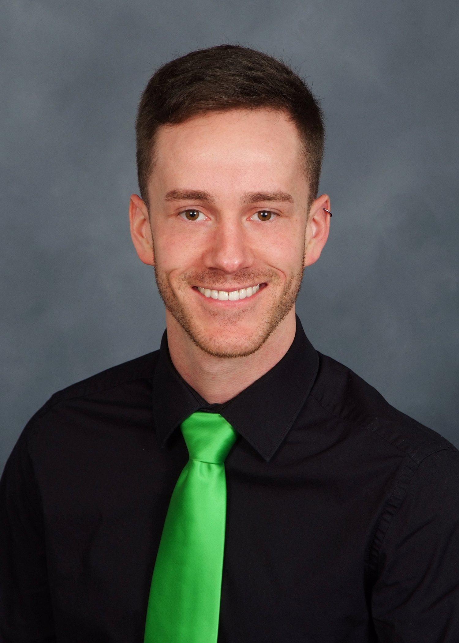 Smiling young man with short brown hair wearing a black shirt and bright green tie against a gray background.