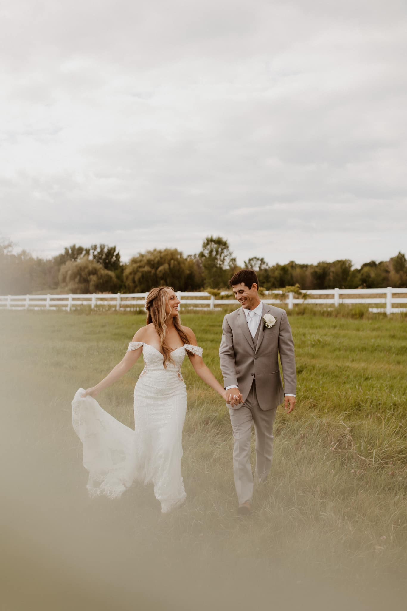 Bride in white off-shoulder gown and groom in light gray suit holding hands and walking on grass with white fence and trees in background.