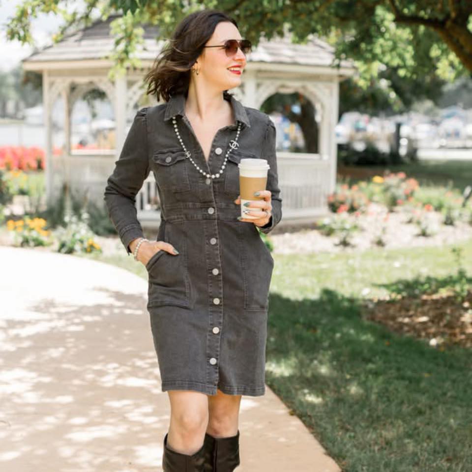 Woman wearing dark buttoned dress and boots, holding coffee cup and walking outside near a white gazebo.