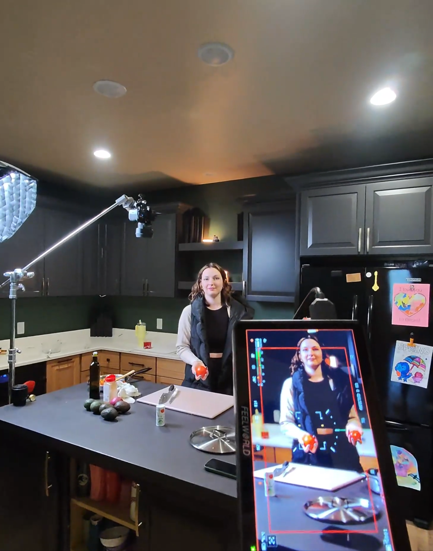 A woman standing in a kitchen holding two tomatoes, with a camera setup filming her and a monitor showing the live recording.