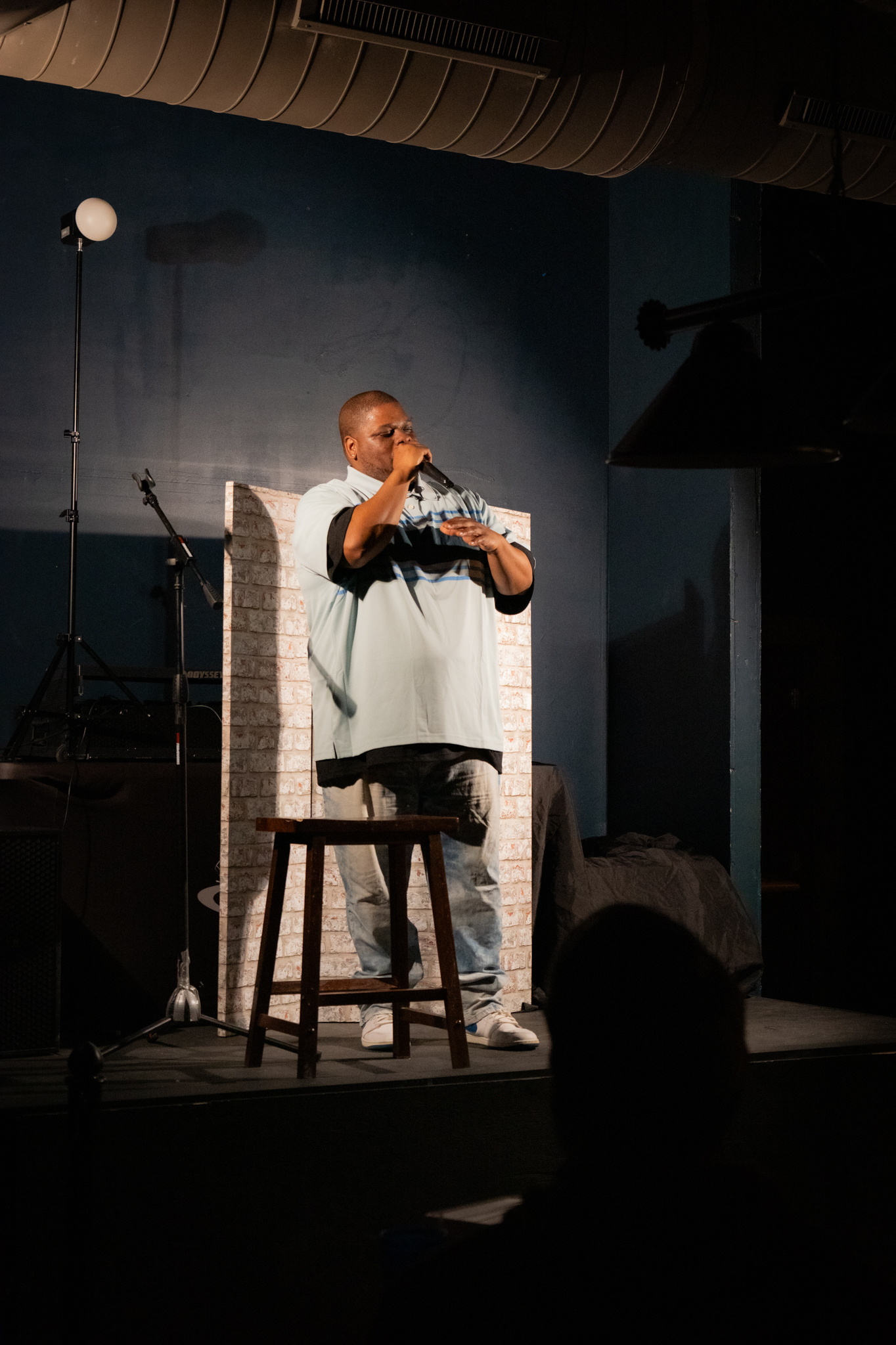 Man in light blue shirt performing stand-up comedy on stage with microphone and stool.
