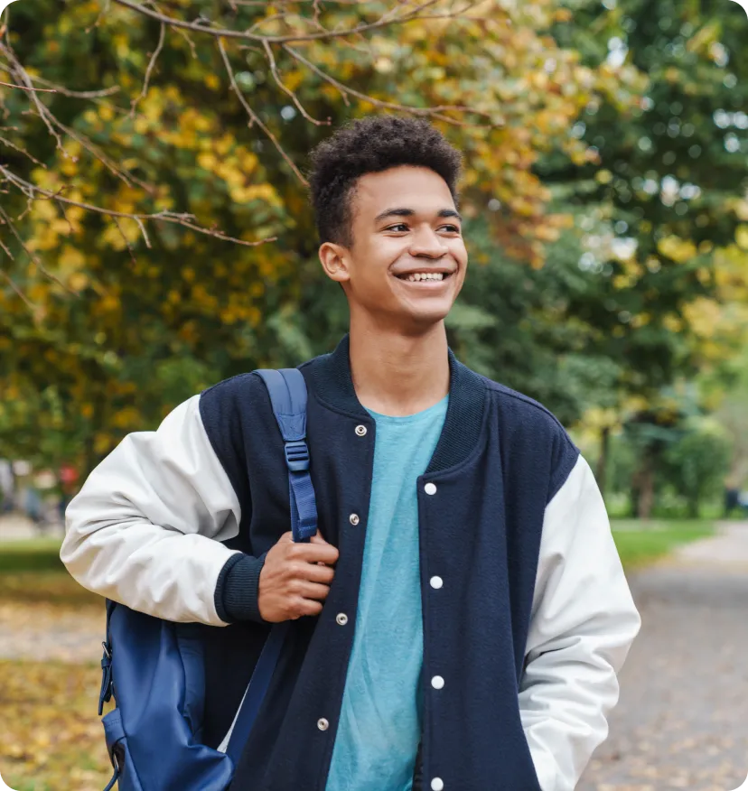 Smiling young man wearing a black and white jacket carrying a blue backpack, standing outdoors with trees in the background.