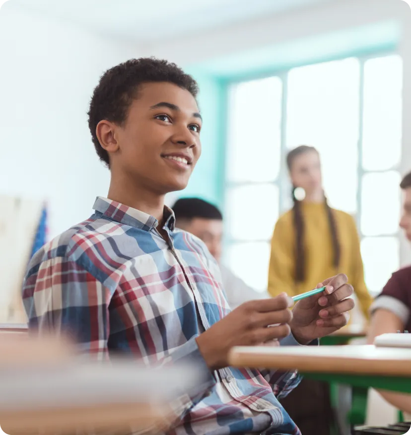 Smiling teenage boy in a plaid shirt holding a pen, sitting in a classroom with other students.