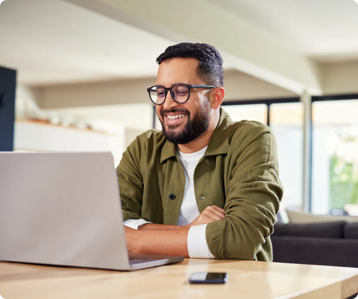 Smiling man wearing glasses and green jacket using a laptop at a wooden table in a bright room.