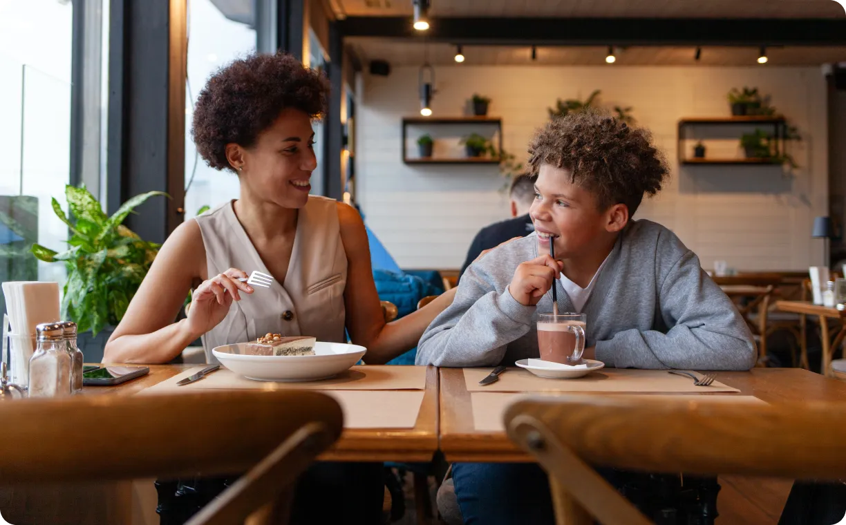 Smiling woman and boy sitting at a cafe table with cake and a hot drink, engaging in a friendly conversation.